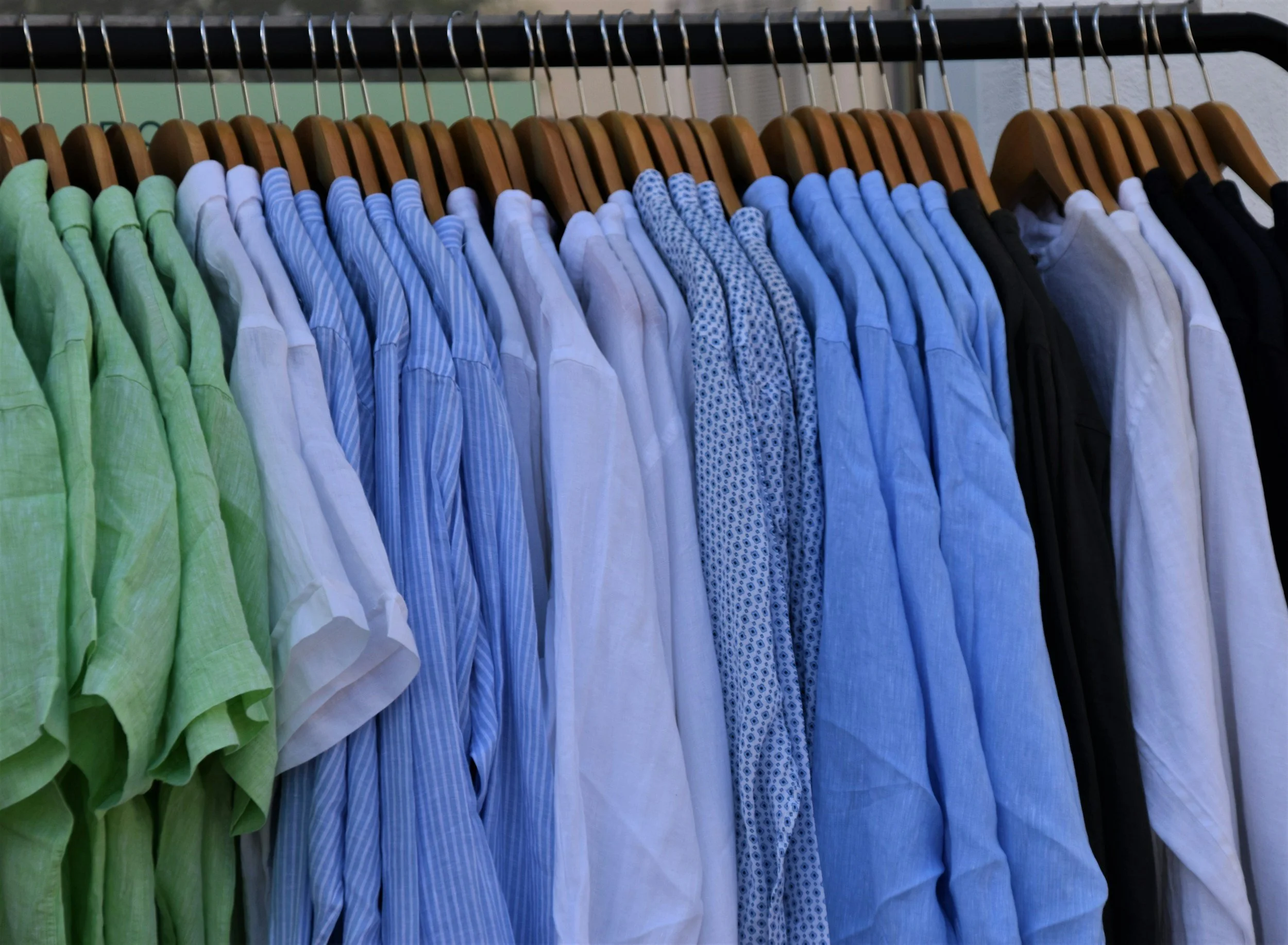 A clothing rack with various men's button-up shirts hanging on wooden hangers, arranged by color from green to black.
