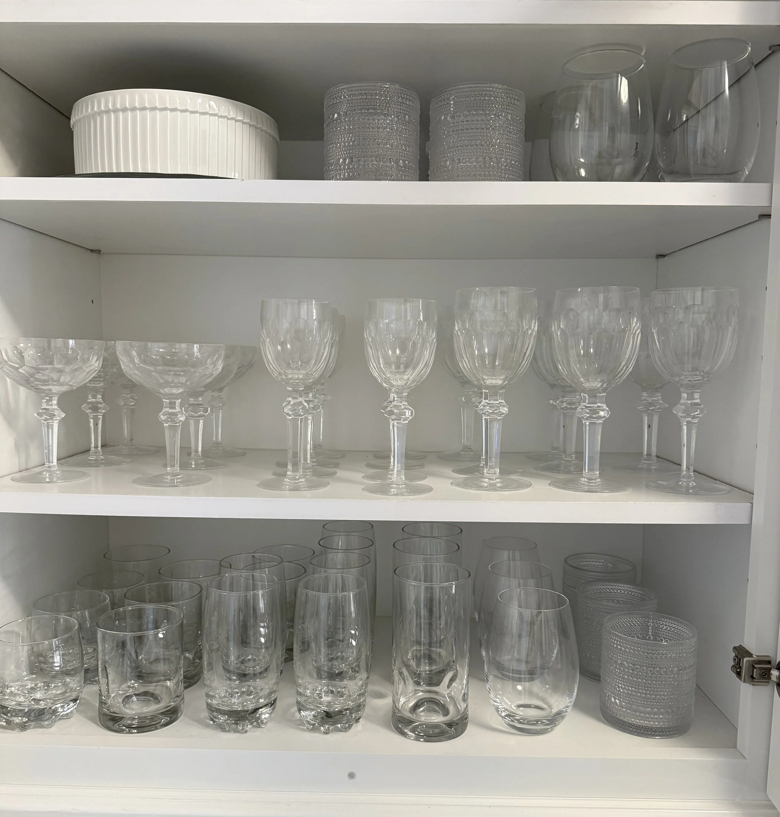 White kitchen cabinet shelves holding various glassware and containers, including wine glasses, tumblers, and small glass jars.