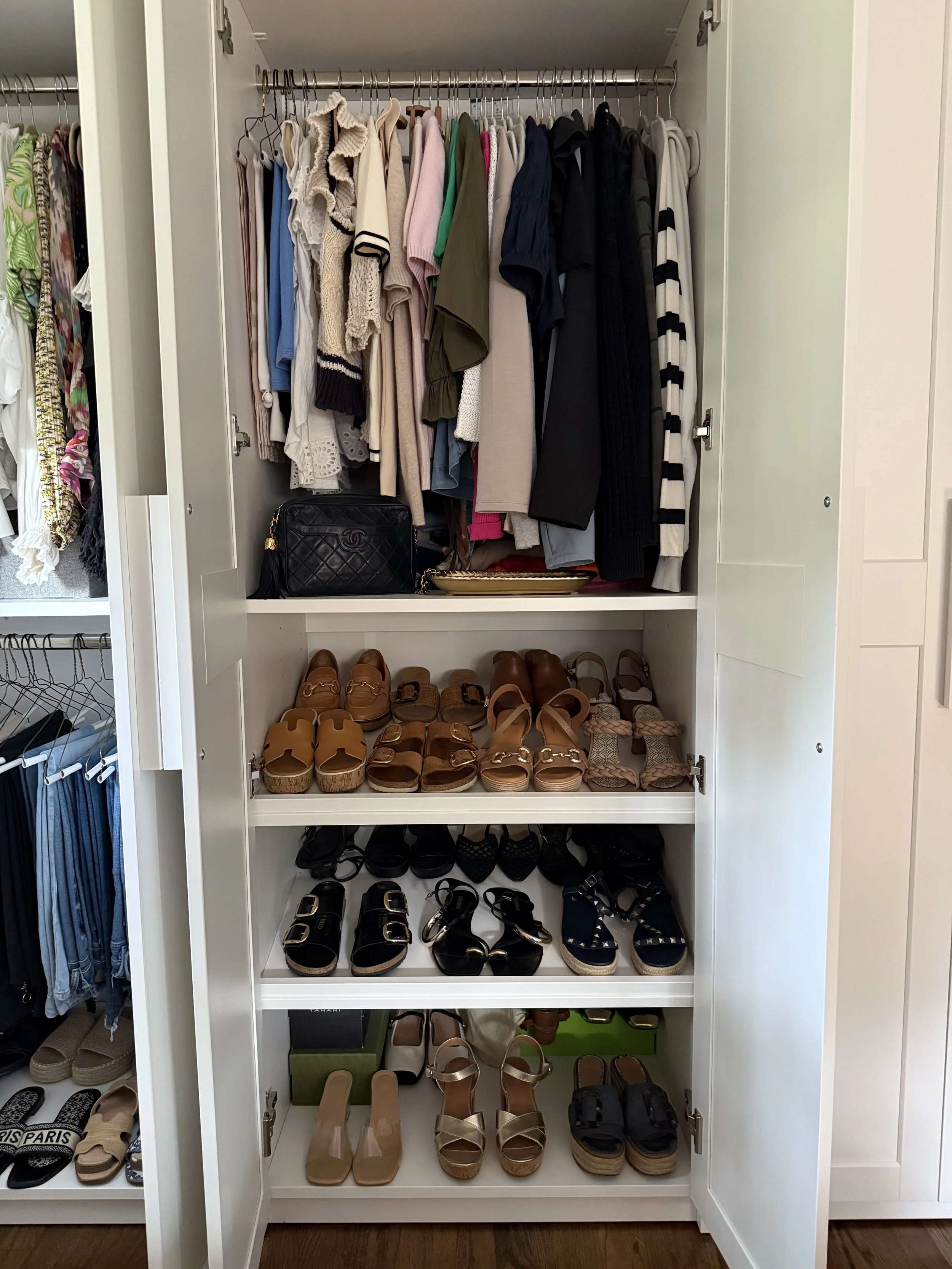 Open wardrobe with hanging clothes on top shelf, middle shelf has various sandals, next shelf has black and tan shoes, bottom shelf has beige heels, tan sandals, and black sandals.