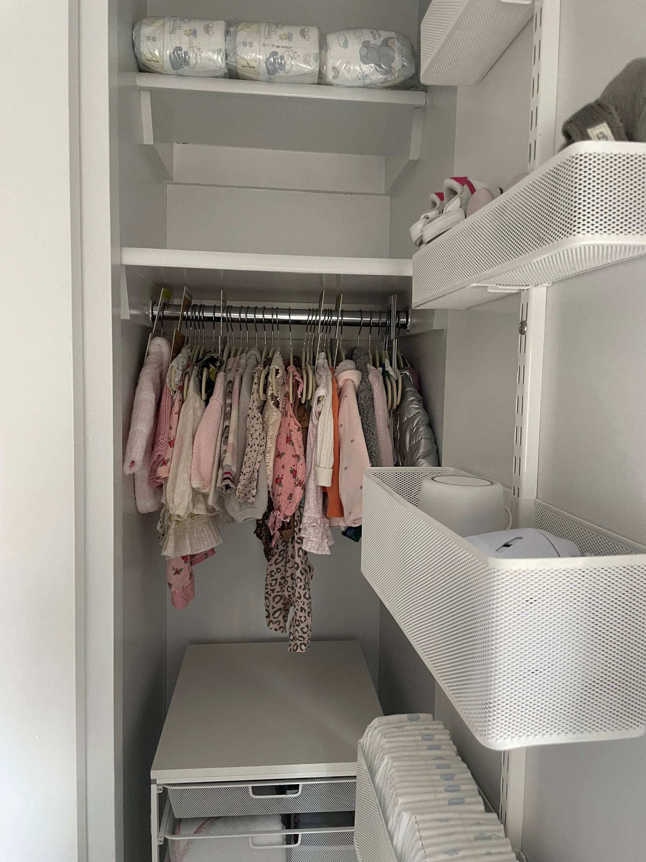A closet with hanging children's clothes, shelves with folded diapers at the top, and storage baskets on the side with baby wipes and other items.