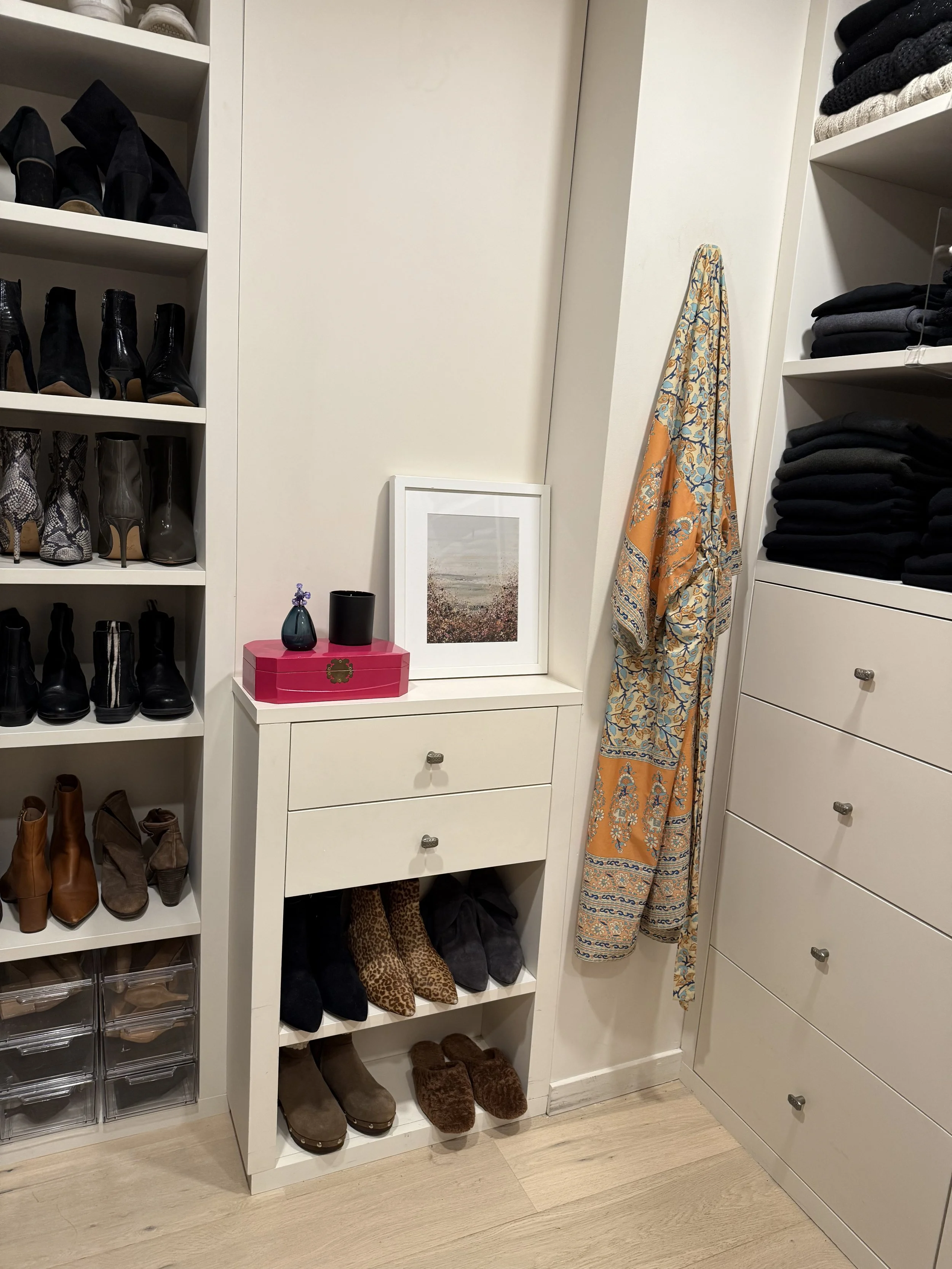 Closet with shelves filled with shoes, a small white table with drawers, a framed picture, decorative items, a patterned scarf hanging on the wall, and folded clothes on shelves.