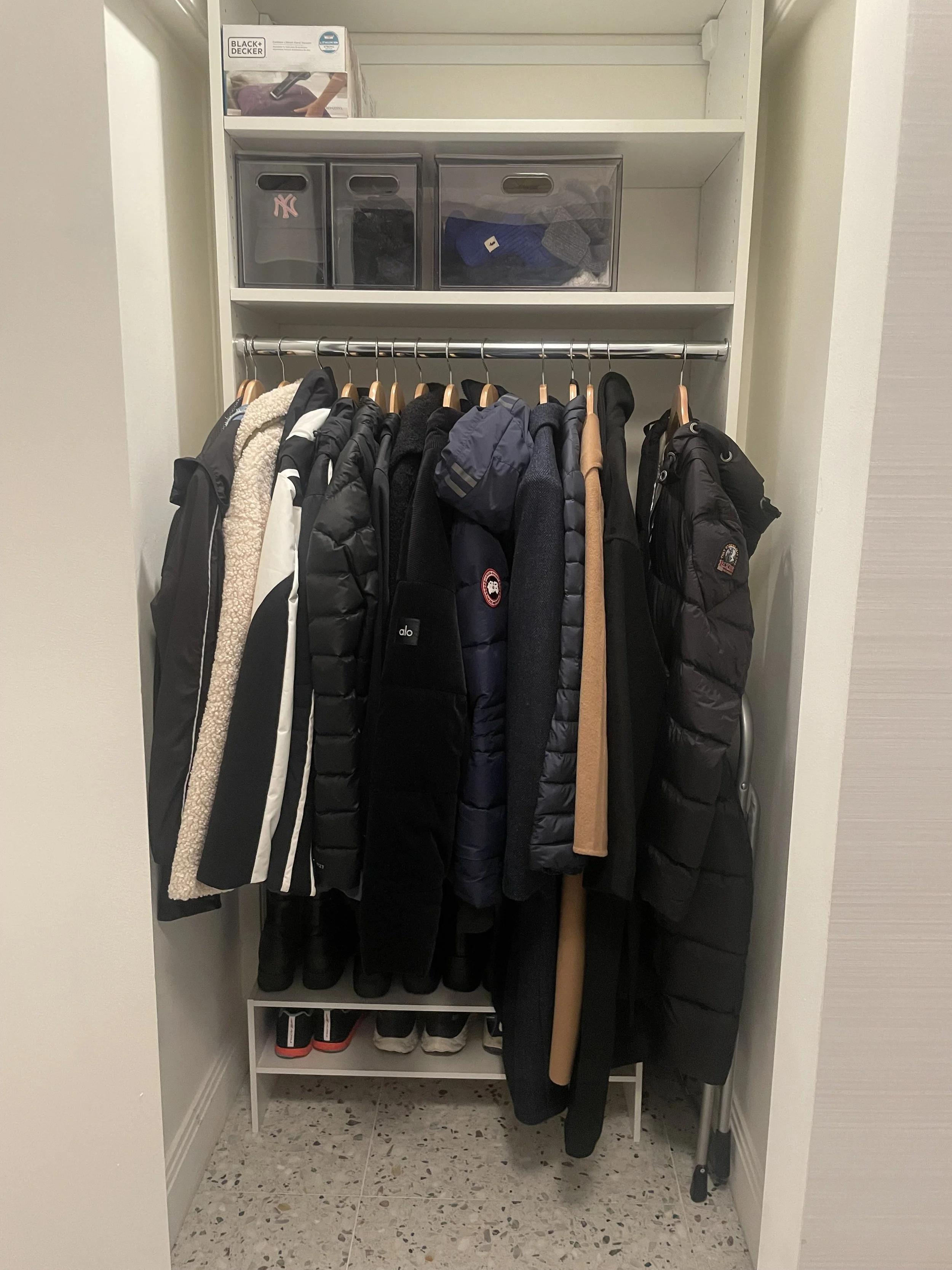 Closet with hanging jackets, coats, and shoes on a white shoe rack at the bottom, plastic storage bins on the shelf above, and a Black & Decker box on the top shelf.