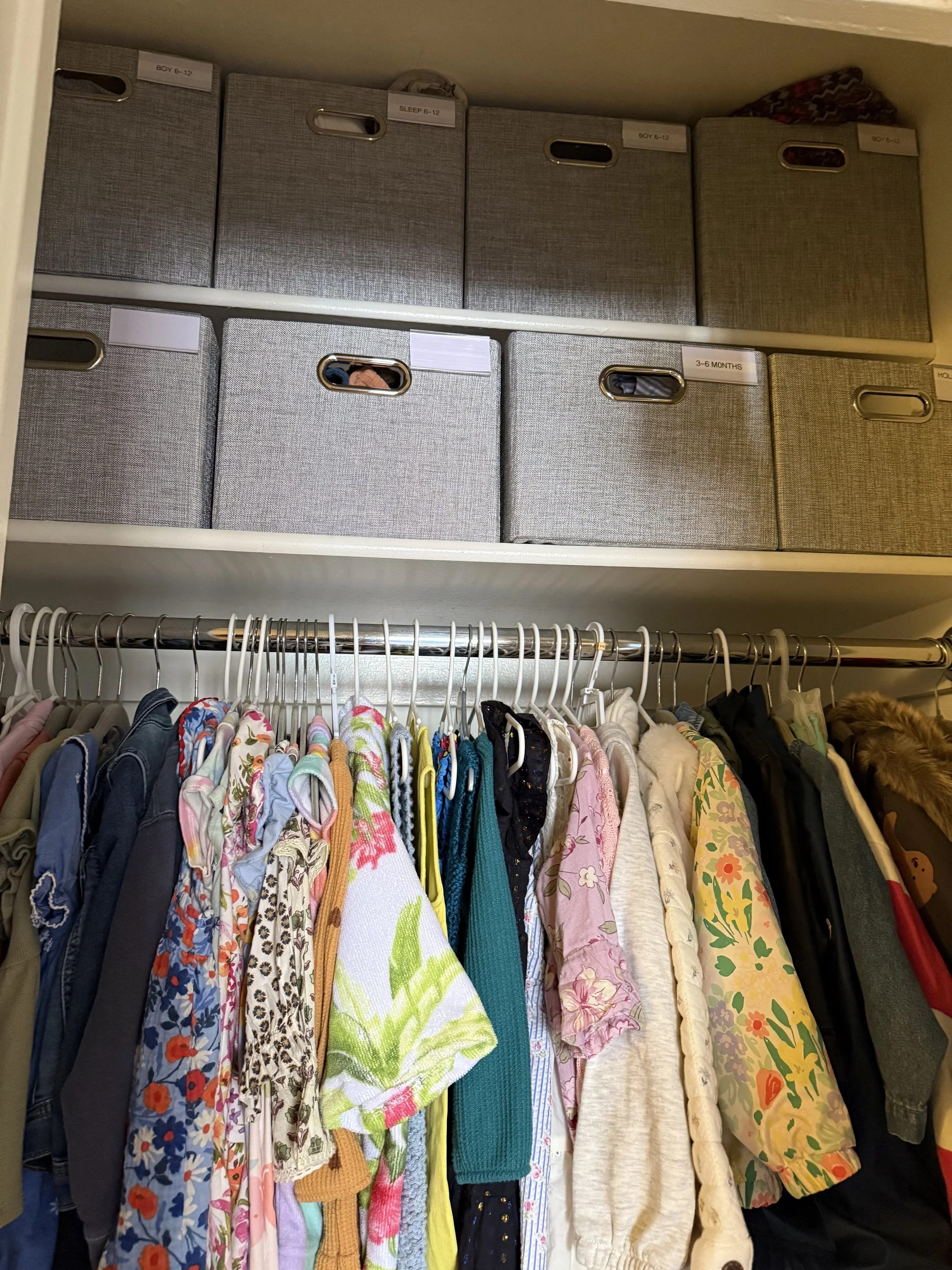 Closet with a row of hanging clothes including jackets, dresses, and shirts, and shelves above filled with gray storage boxes labeled with age ranges and categories.