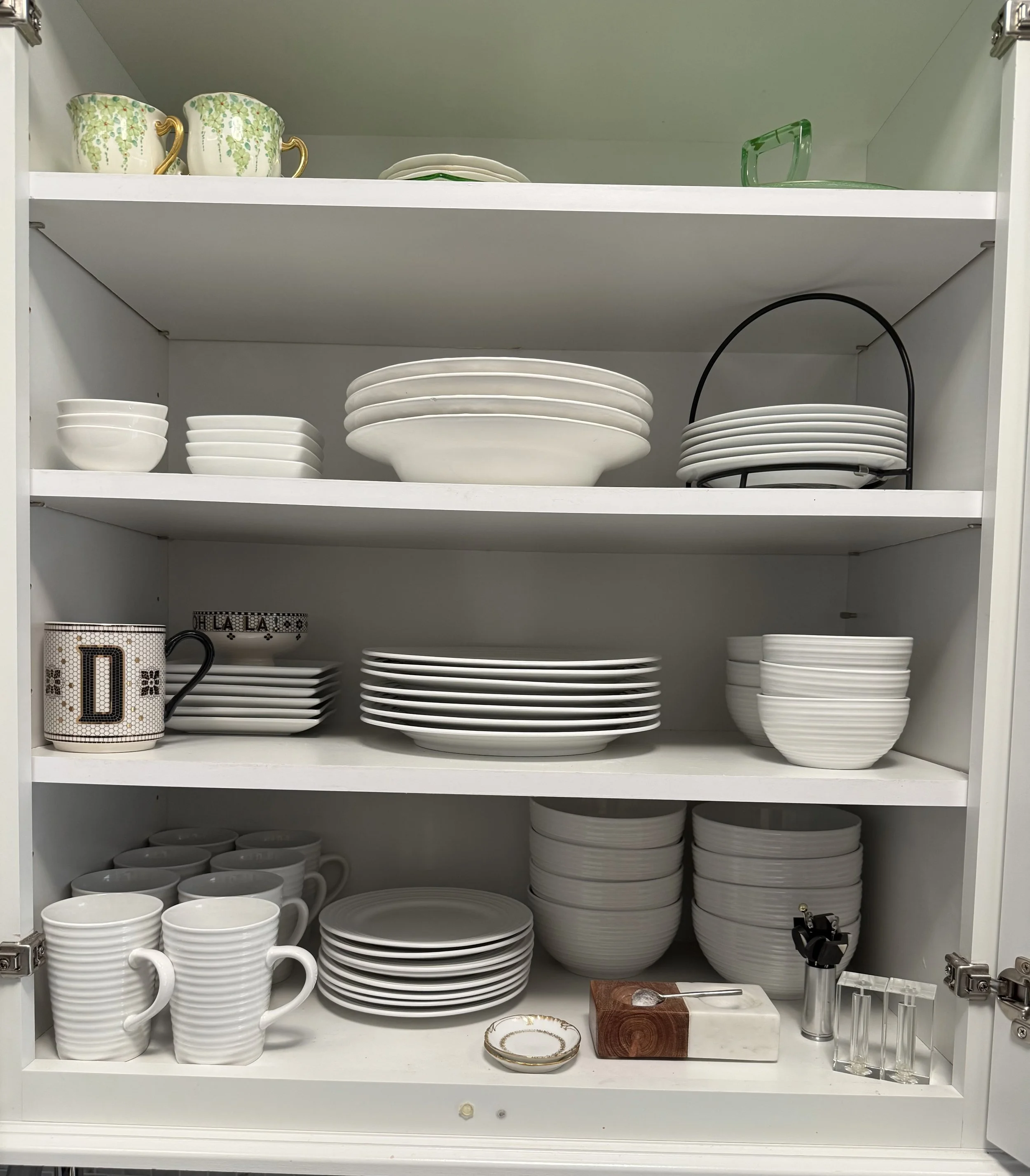 Open cabinet filled with white dinnerware including bowls, plates, and mugs, with some decorative items, and a cutting board with a small plate on it.