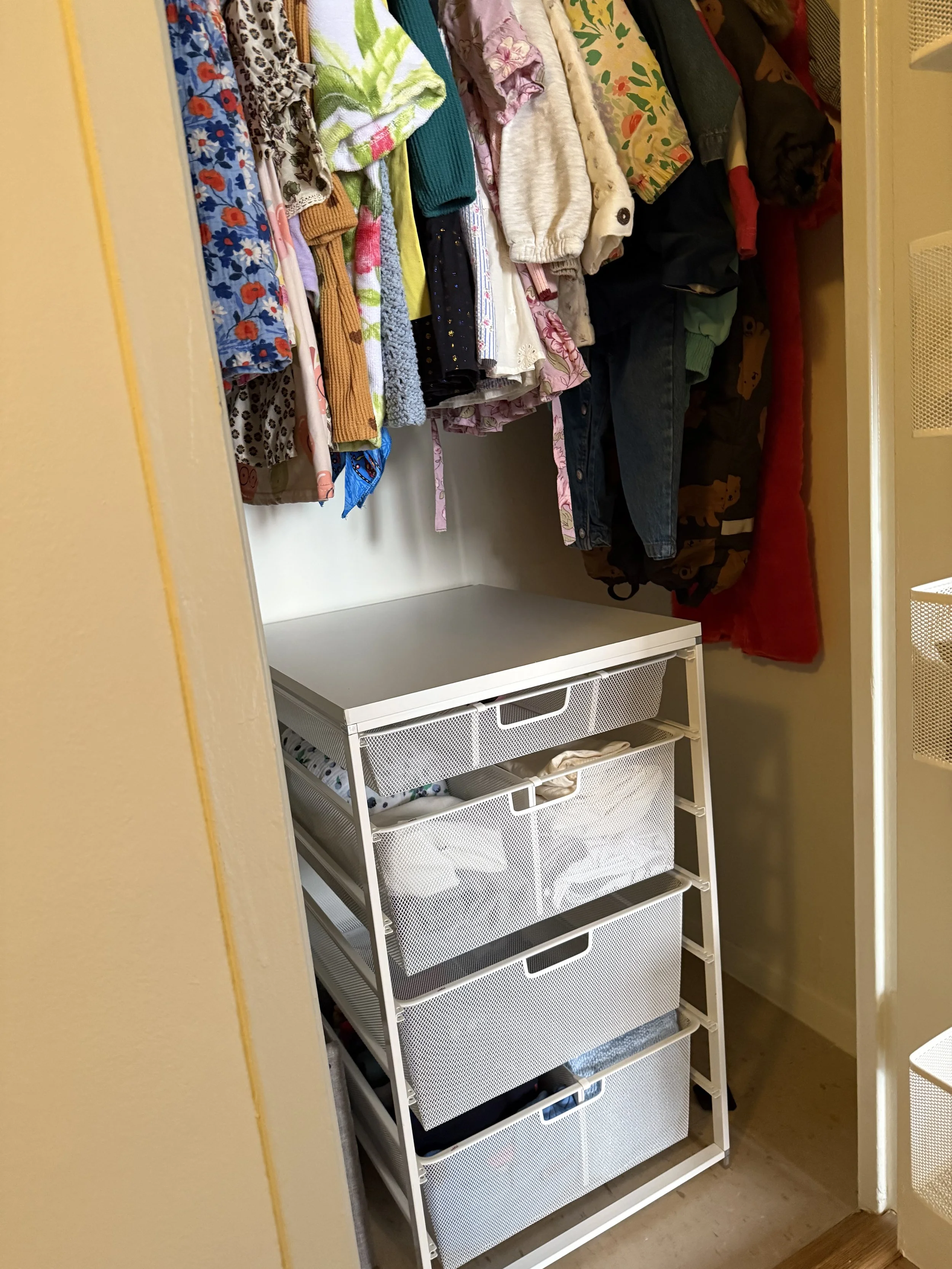 A small closet with colorful clothes hanging on a rod and a white storage unit with four mesh drawers at the bottom right.