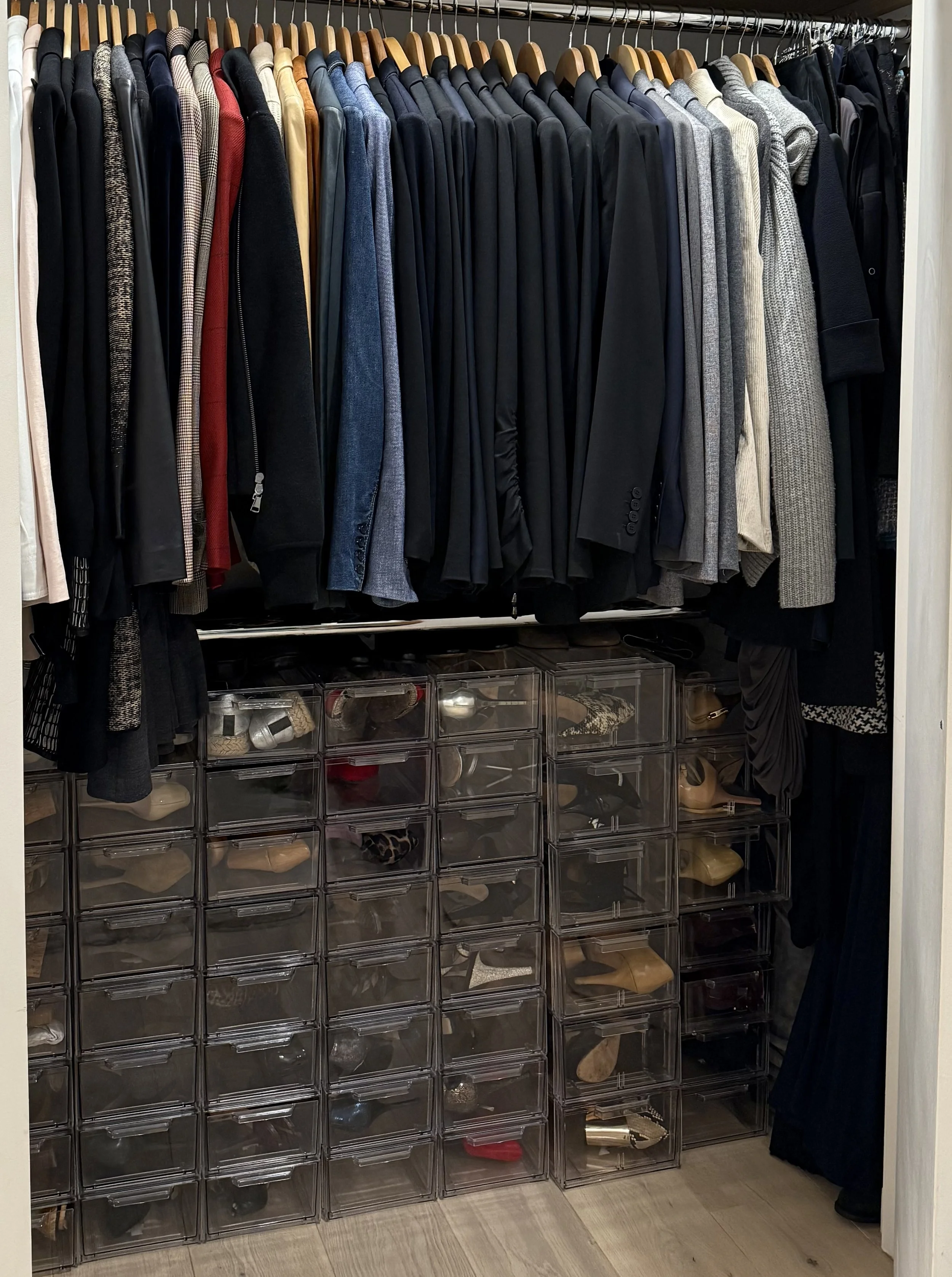 A closet with a row of men's suits and jackets hanging on wooden hangers above multiple transparent plastic shoe storage drawers filled with various shoes.