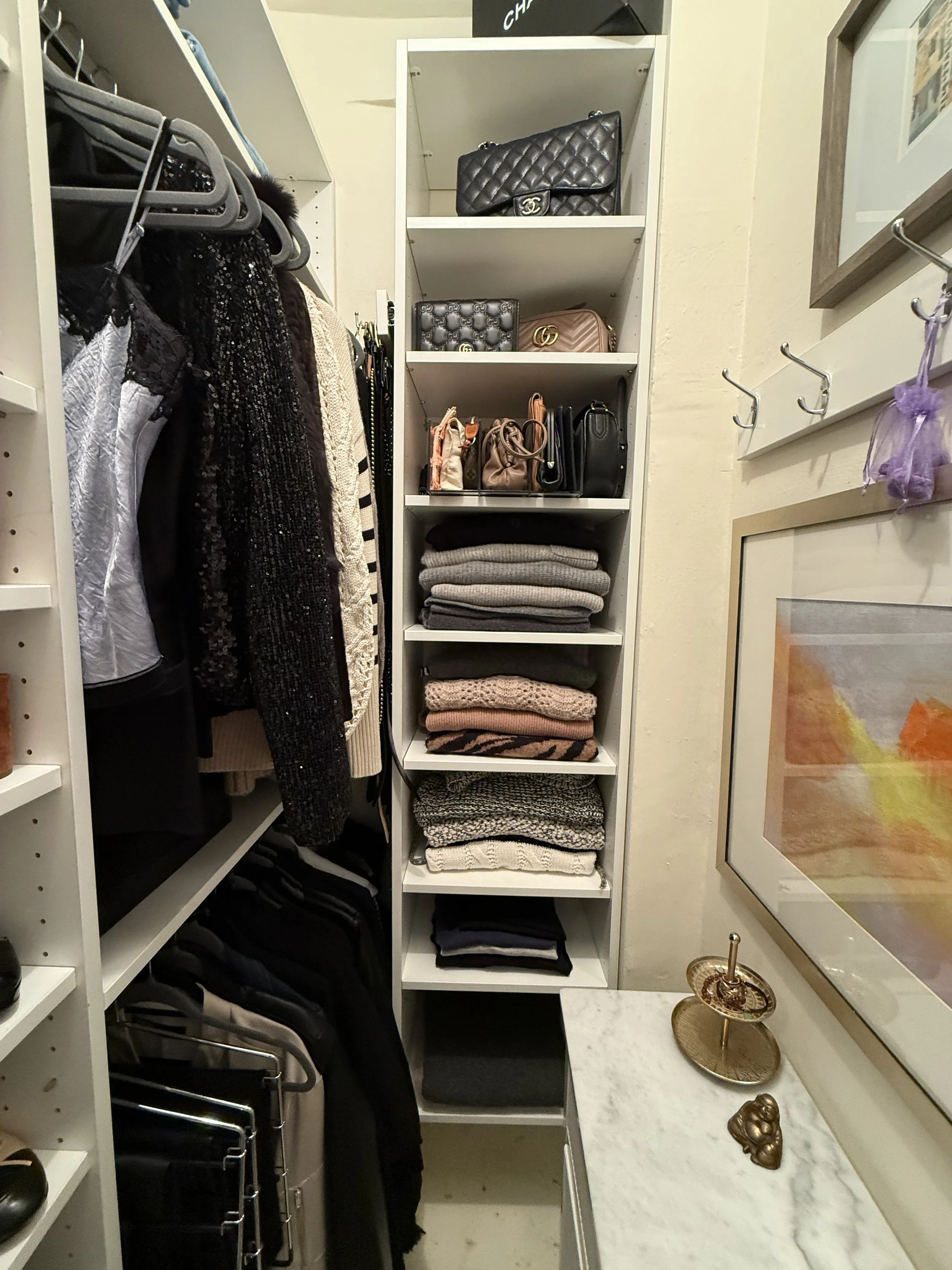 Closet with clothing, handbags, and folded sweaters on white shelving and hanging racks, next to a marble-topped cabinet with decorative items and framed art.