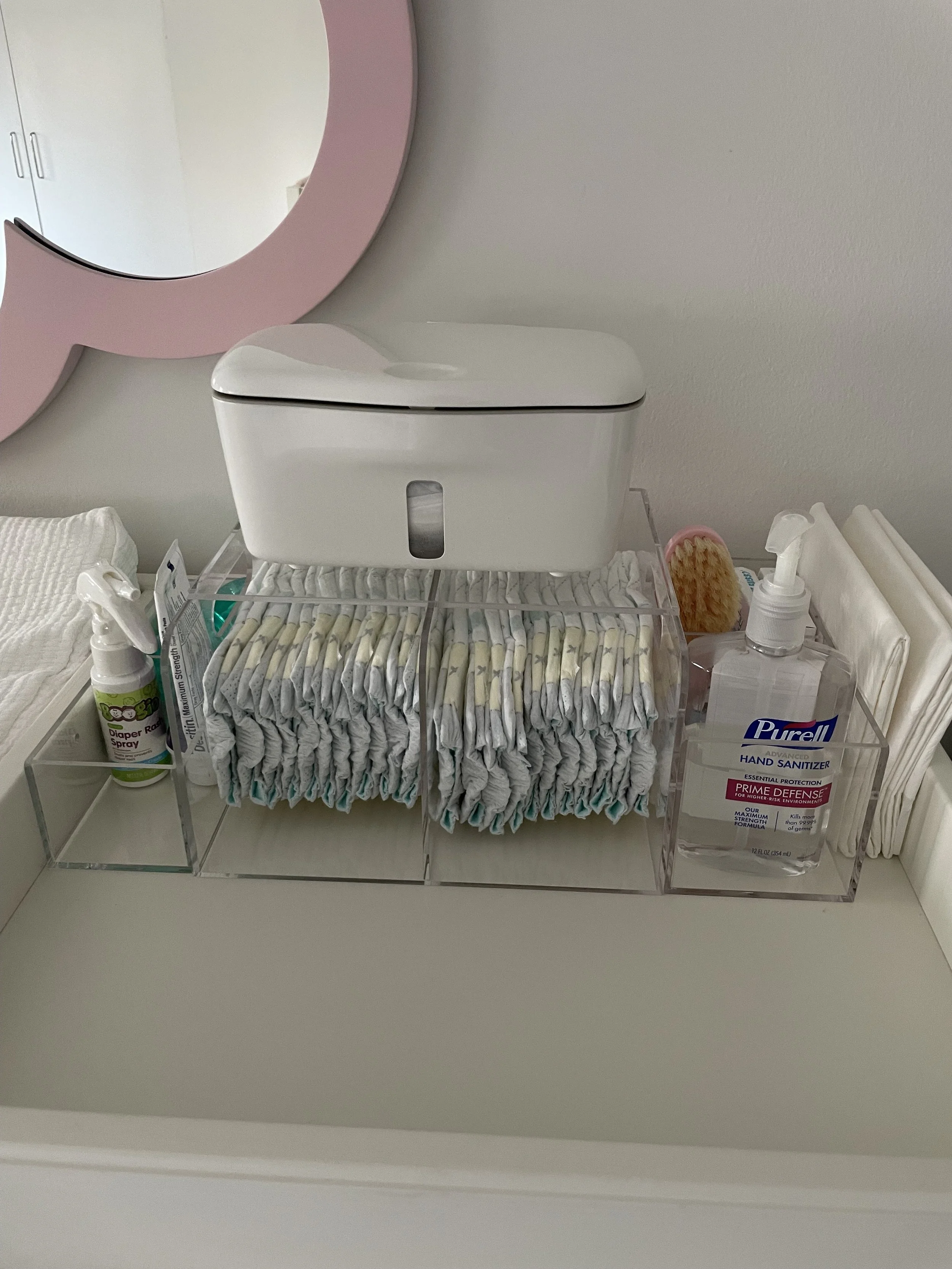 A white countertop with a diaper pale, hand sanitizer, a container of wipes, and a baby brush, all organized in a clear acrylic holder, with a round pink mirror on the wall behind.