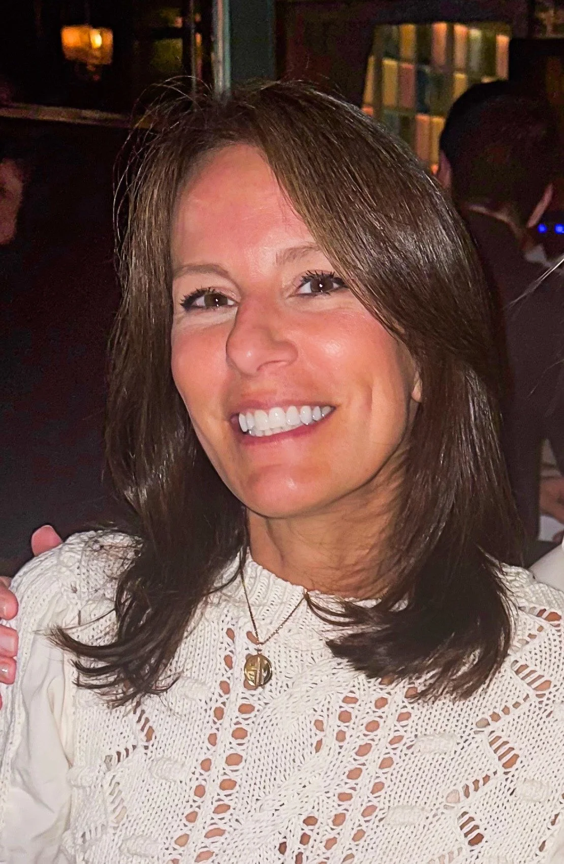 A woman with shoulder-length brown hair, smiling, wearing a white crochet top and a small gold pendant necklace, in a dimly lit indoor setting.