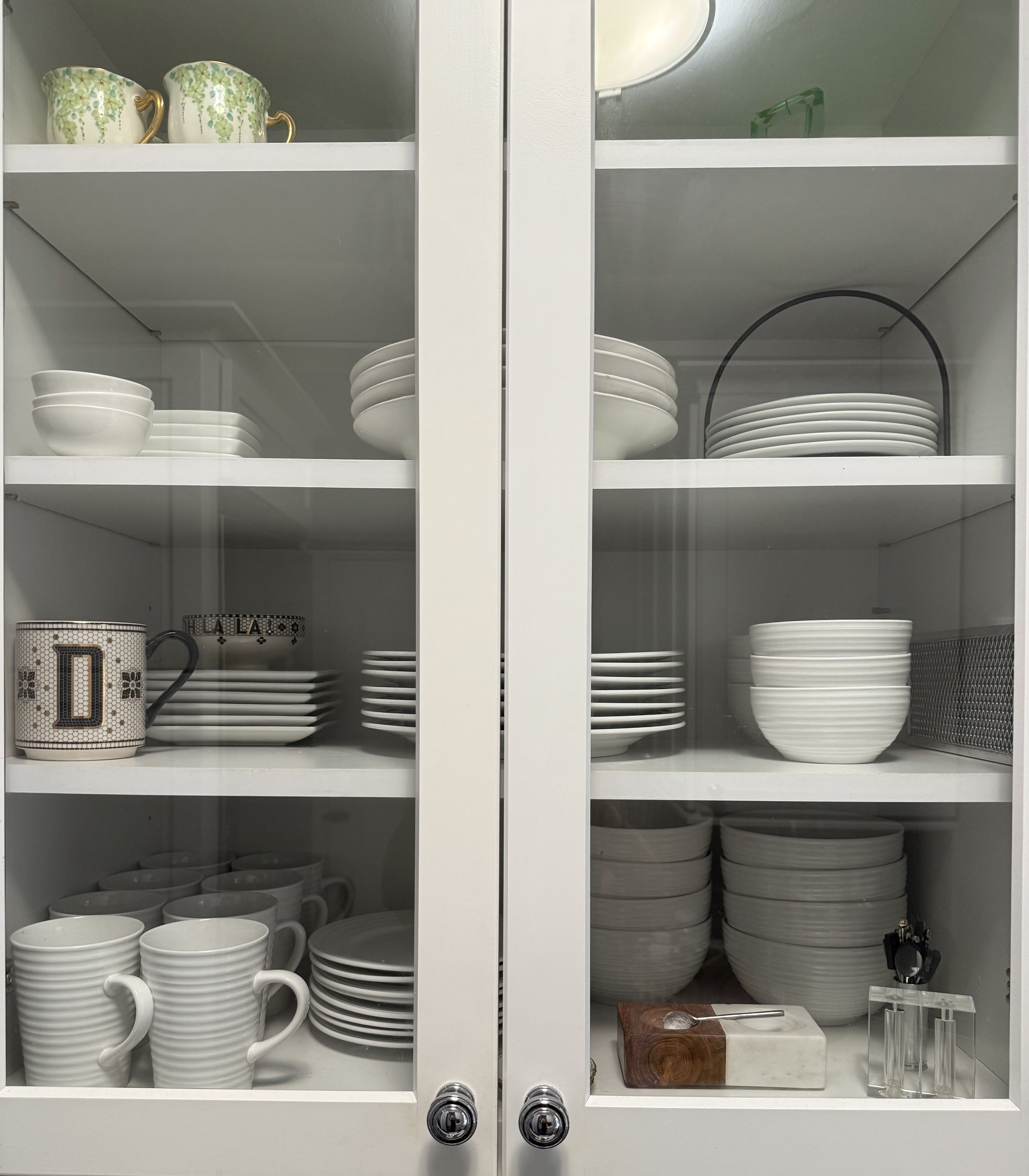A white kitchen cabinet with glass doors displaying white dishes, bowls, mugs, and a decorative mug with a letter 'D'. Items are organized on multiple shelves, with some bowls stacked and a wooden serving board and glass salt and pepper shakers on th