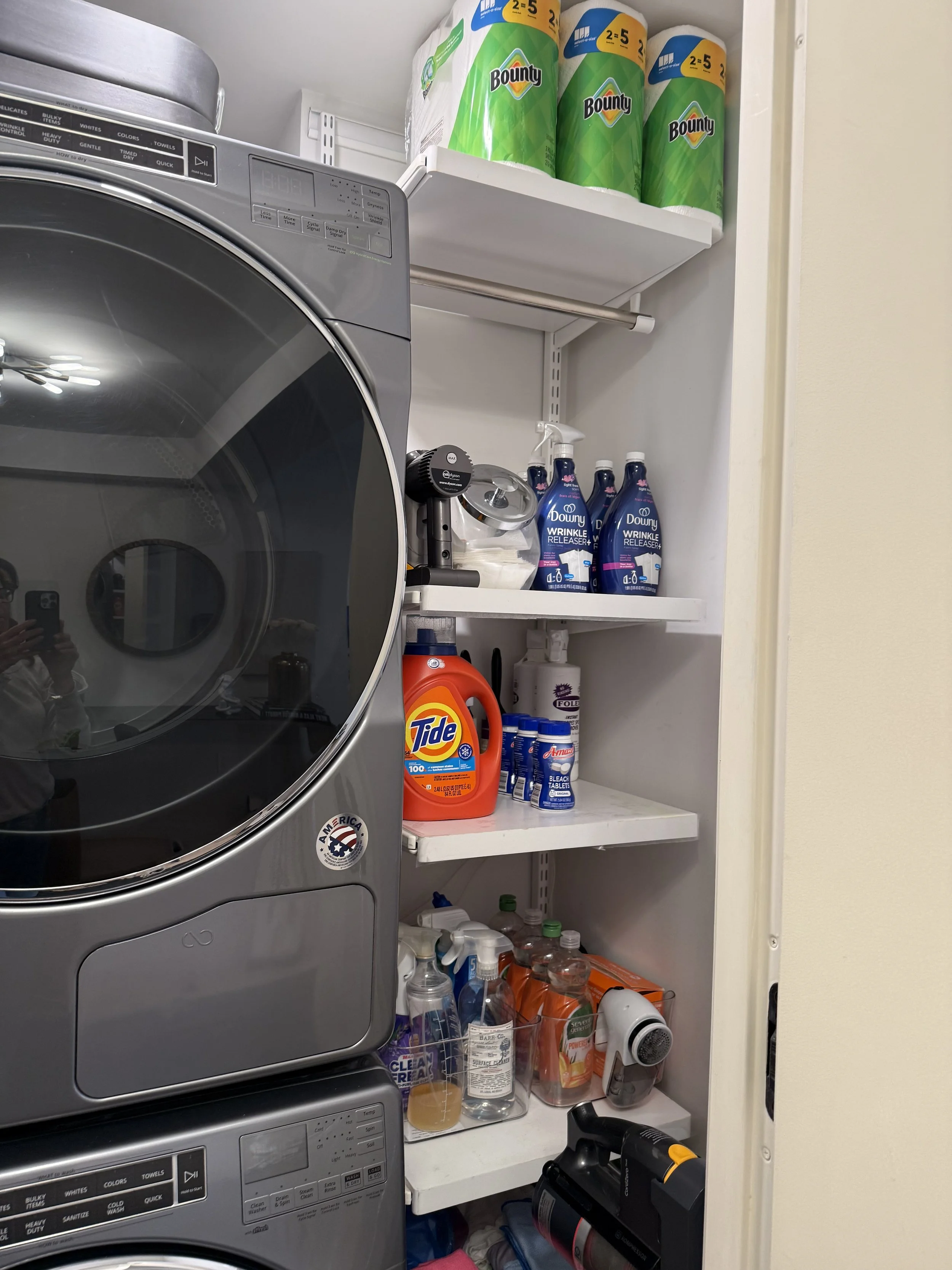 Laundry room with shelves containing containers of Bounty paper towels, Downy fabric softener, Tide detergent, disinfectant spray, and various cleaning supplies.