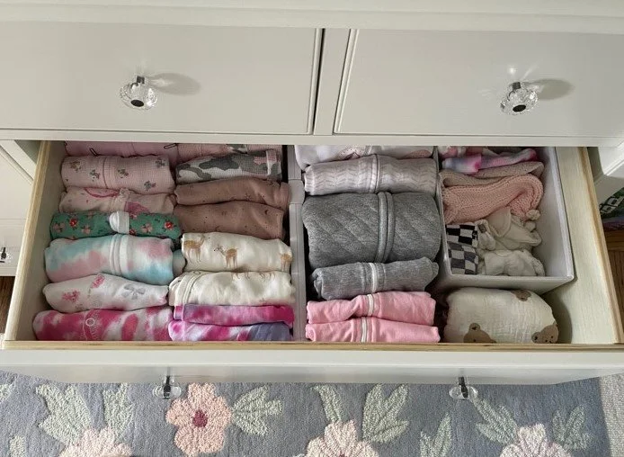 Open drawer with neatly folded baby clothes arranged by color and type, including onesies, socks, and blankets.