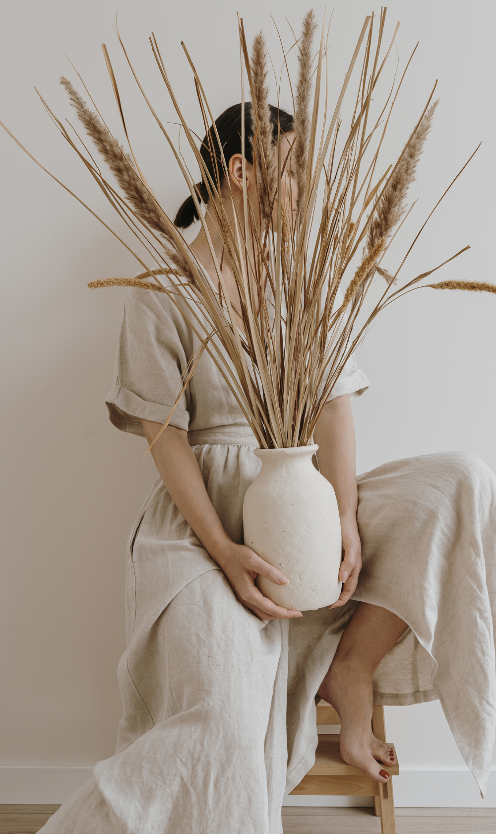 Person sitting on a stool holding a large beige ceramic vase filled with tall dried pampas grass, wearing a beige linen dress, with a plain white wall background. Austin Aesthetics Club | Medspa in Austin, TX | Botox & Fillers