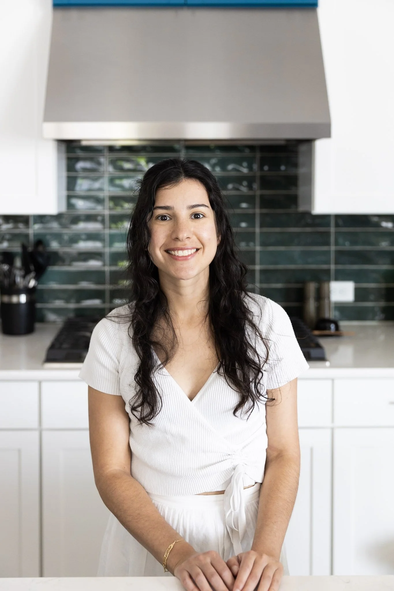 A woman with long dark curly hair smiling in a modern kitchen.