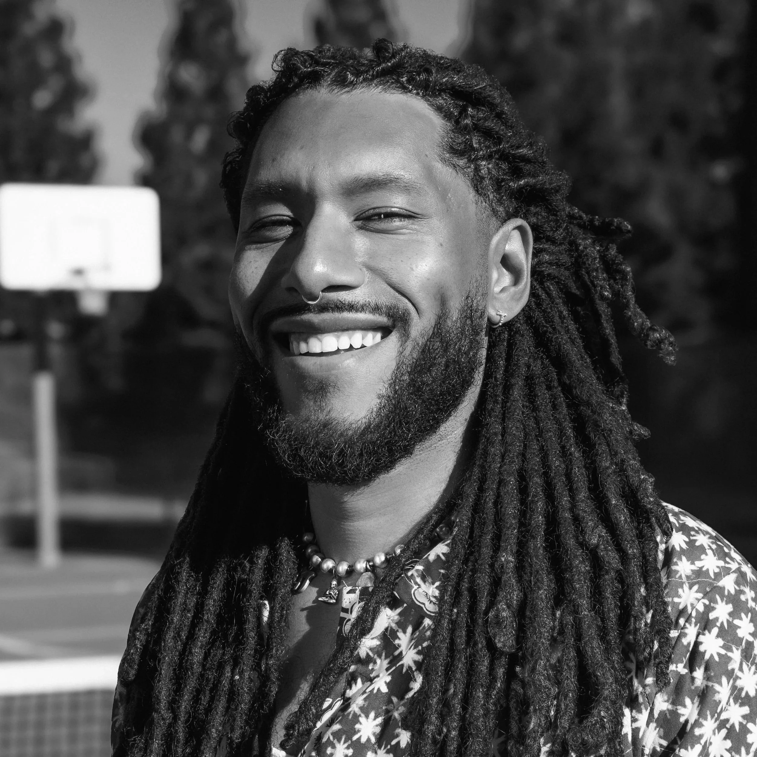 A man with dreadlocks, beard, and earrings smiling outdoors, wearing a patterned shirt and a beaded necklace, with a basketball court and trees in the background.