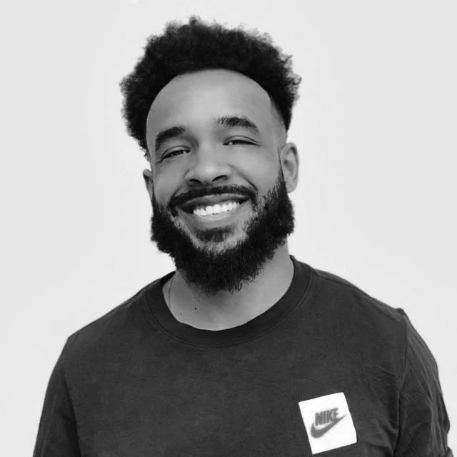 Black and white photo of a smiling man with curly hair and a beard, wearing a Nike T-shirt.