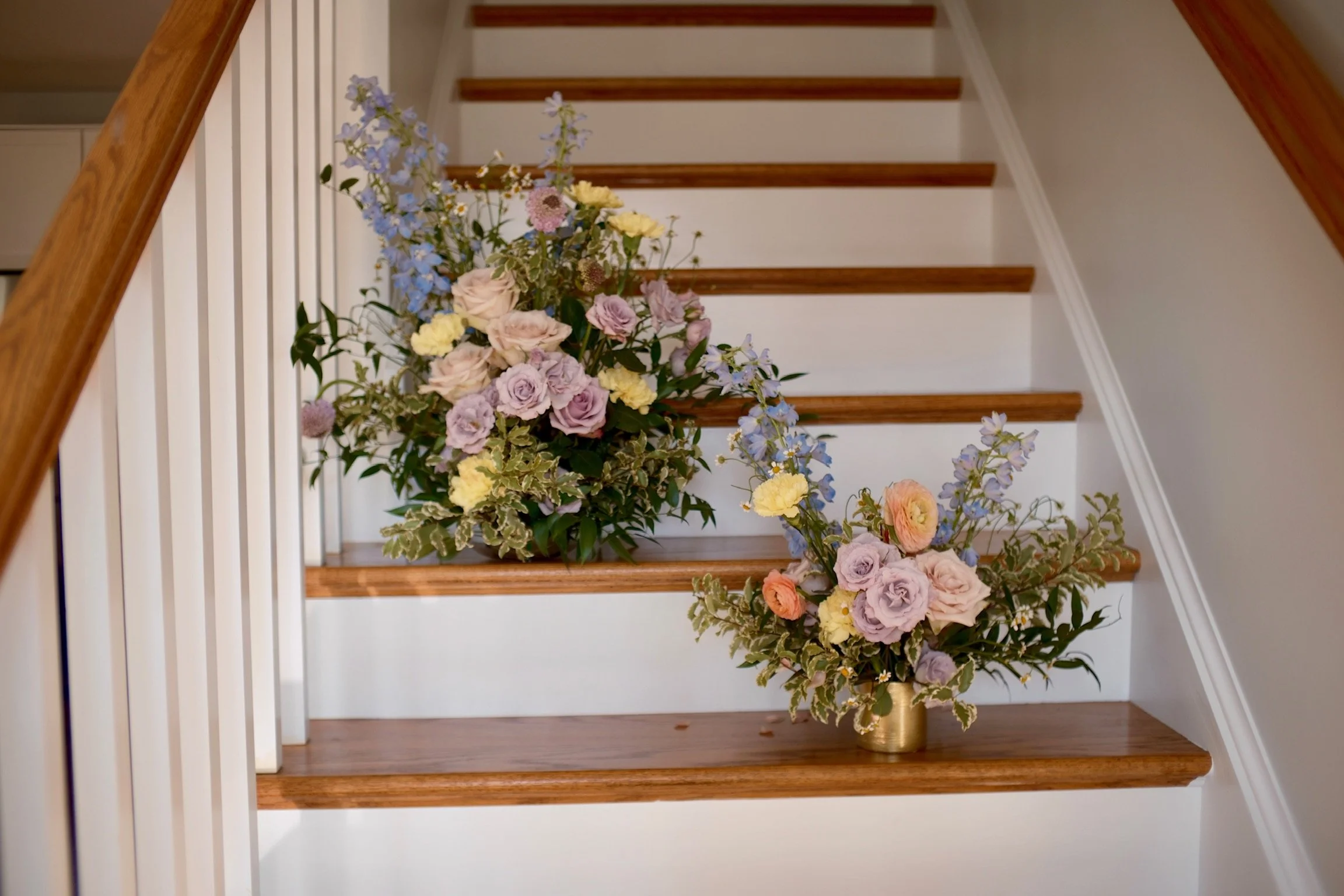 Two large floral arrangements of pastel-colored roses, ranunculus, and delphiniums on a staircase with wooden steps and white risers.