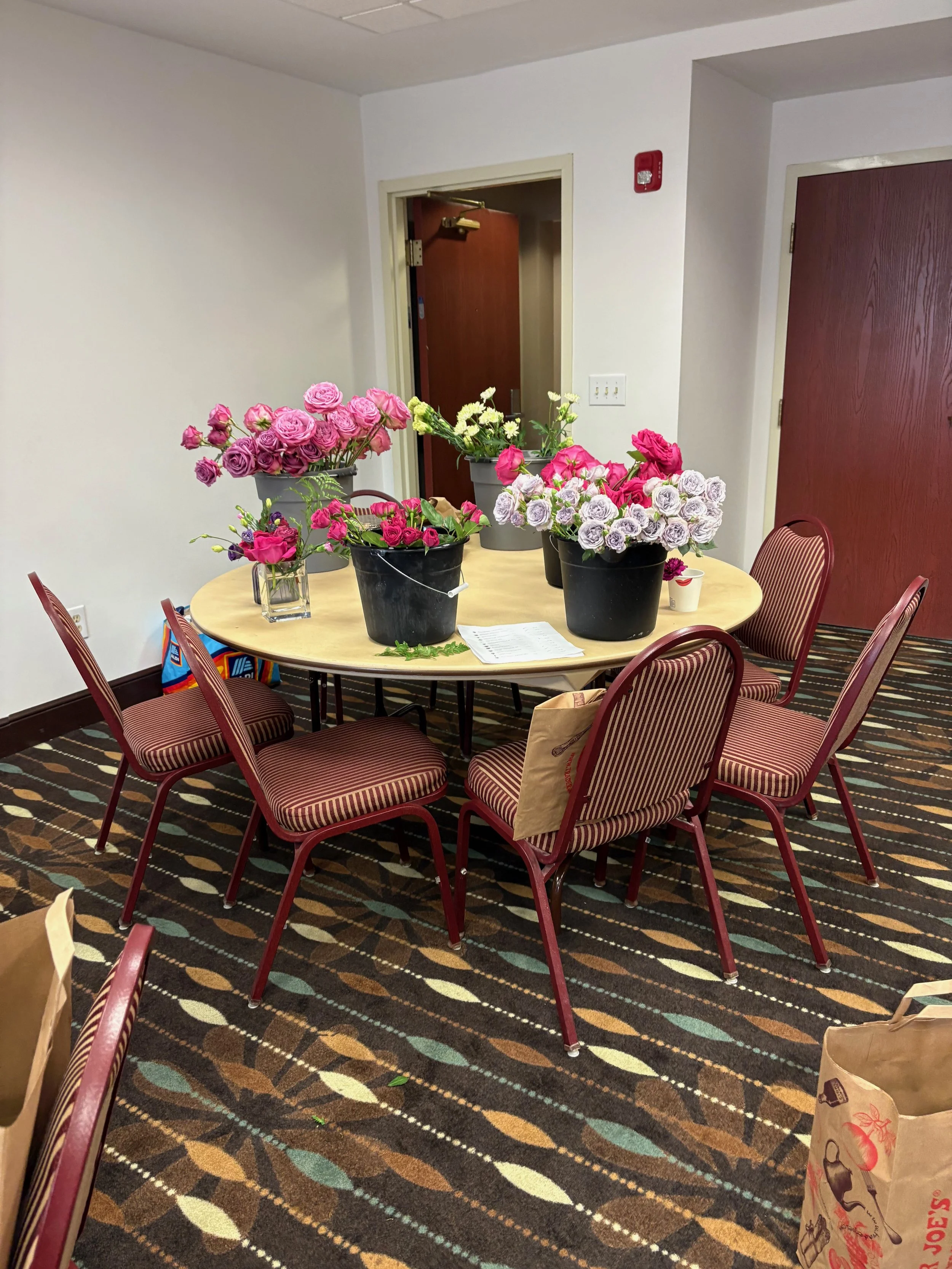 A round table with six red and beige striped chairs around it. The table has various vases with pink, purple, and white flowers. The room has beige walls, one door open, and a patterned carpet.