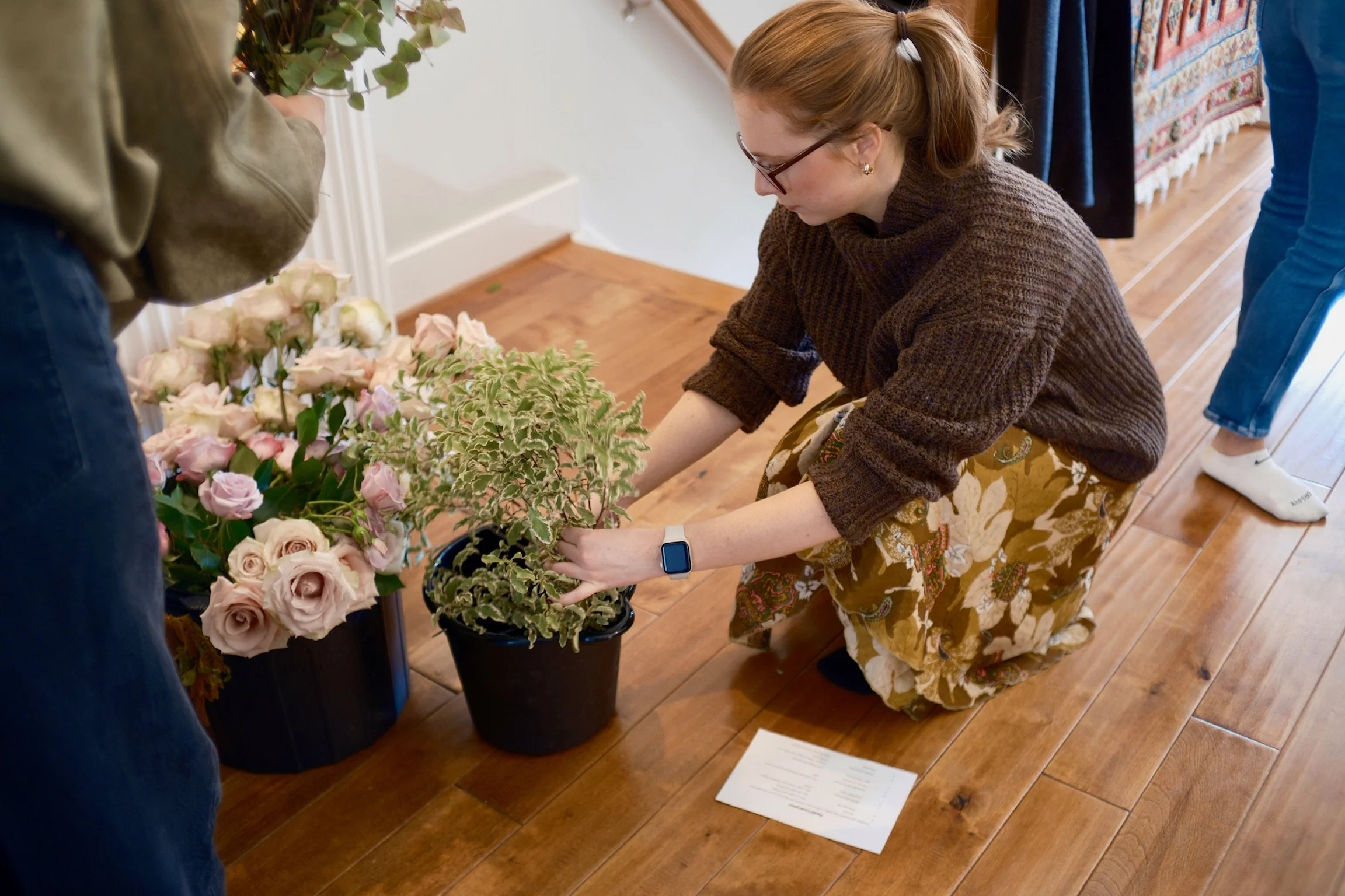 A woman with red hair, glasses, and a smartwatch is kneeling on wooden floor arranging pink and cream roses and green foliage in a black bucket, with another person nearby holding flowers.