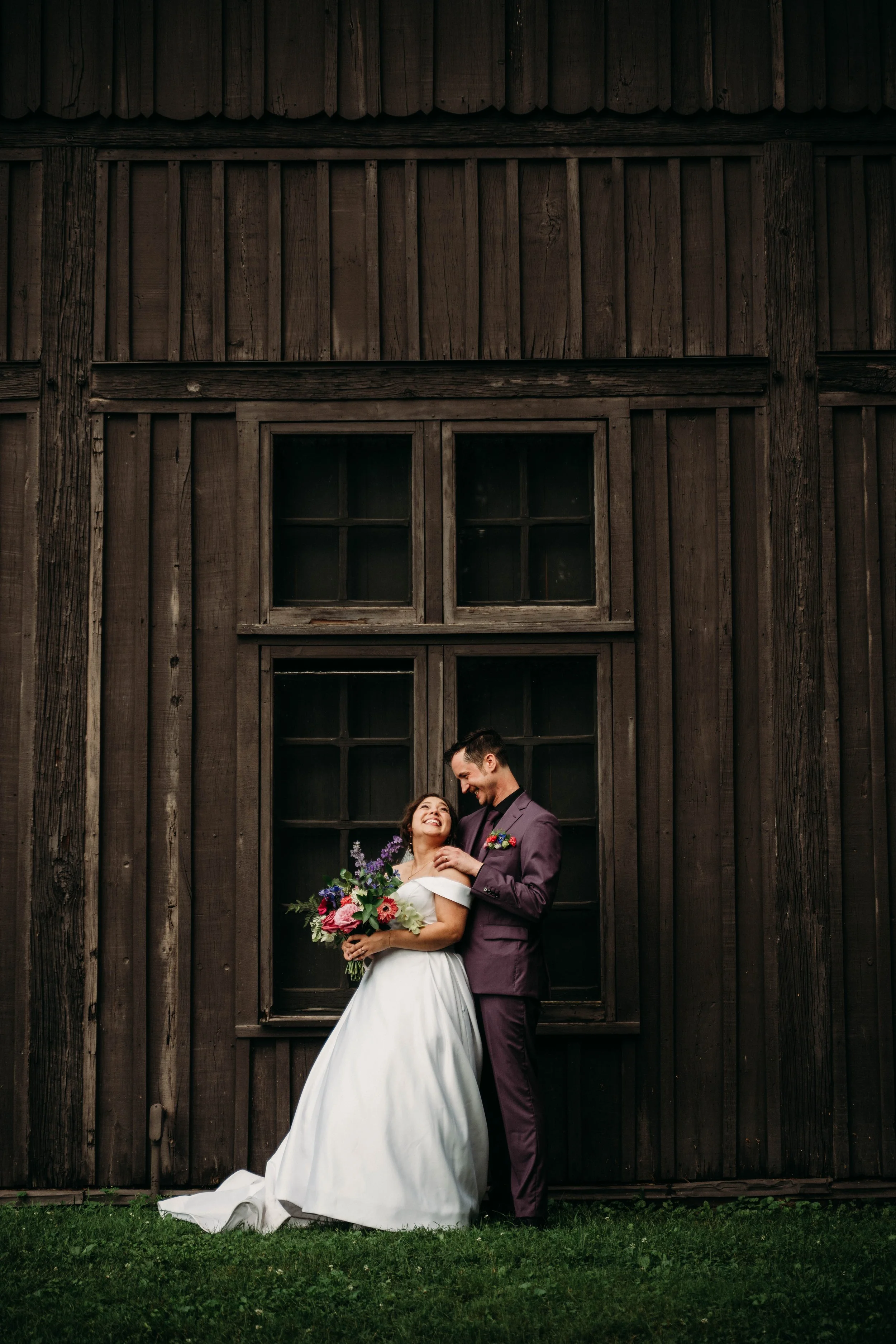 A bride and groom standing in front of a rustic wooden barn, smiling and looking at each other. The bride is holding a bouquet of flowers.