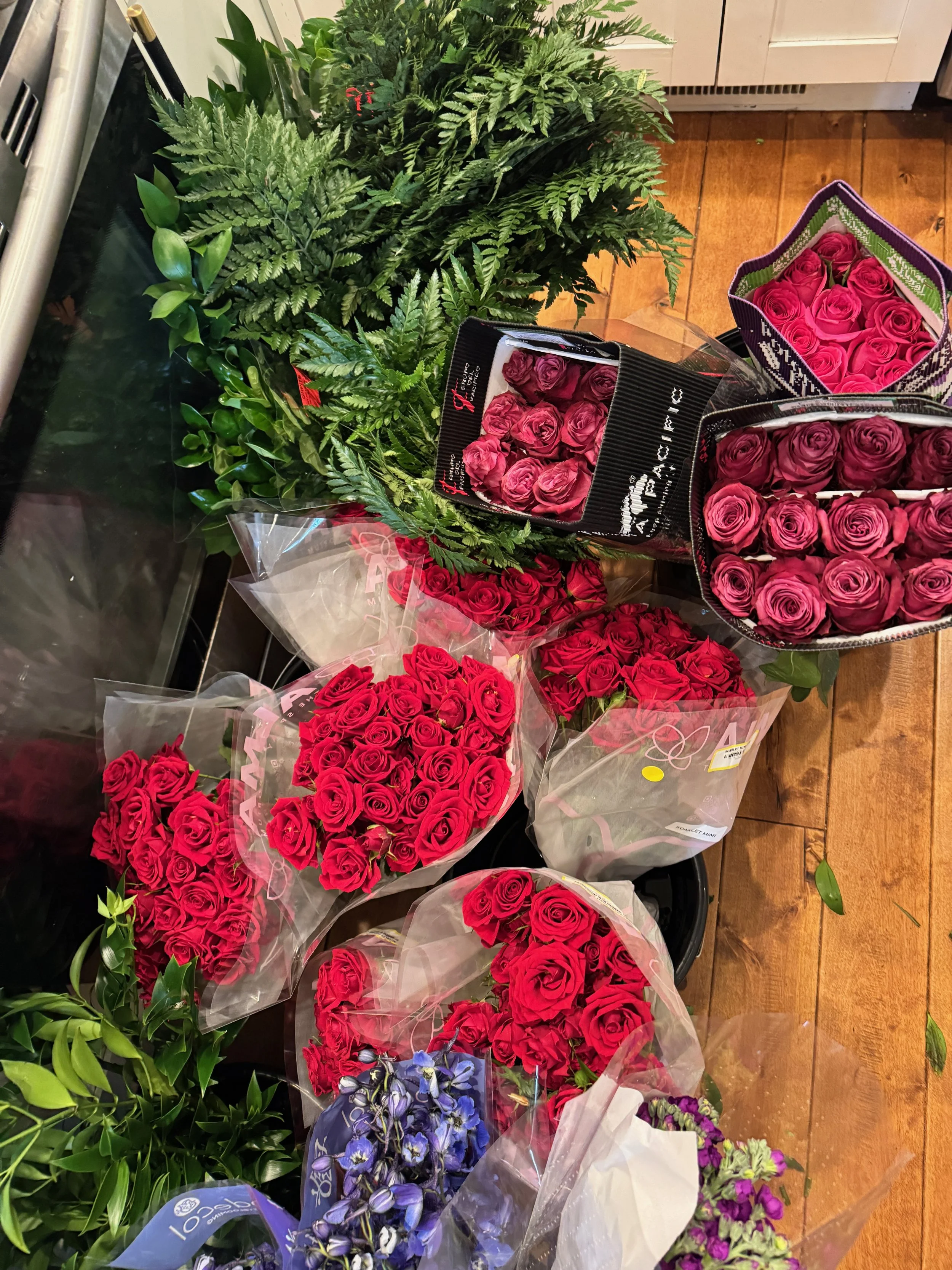 A variety of floral bouquets including red roses, purple flowers, and green foliage arranged on a wooden floor.