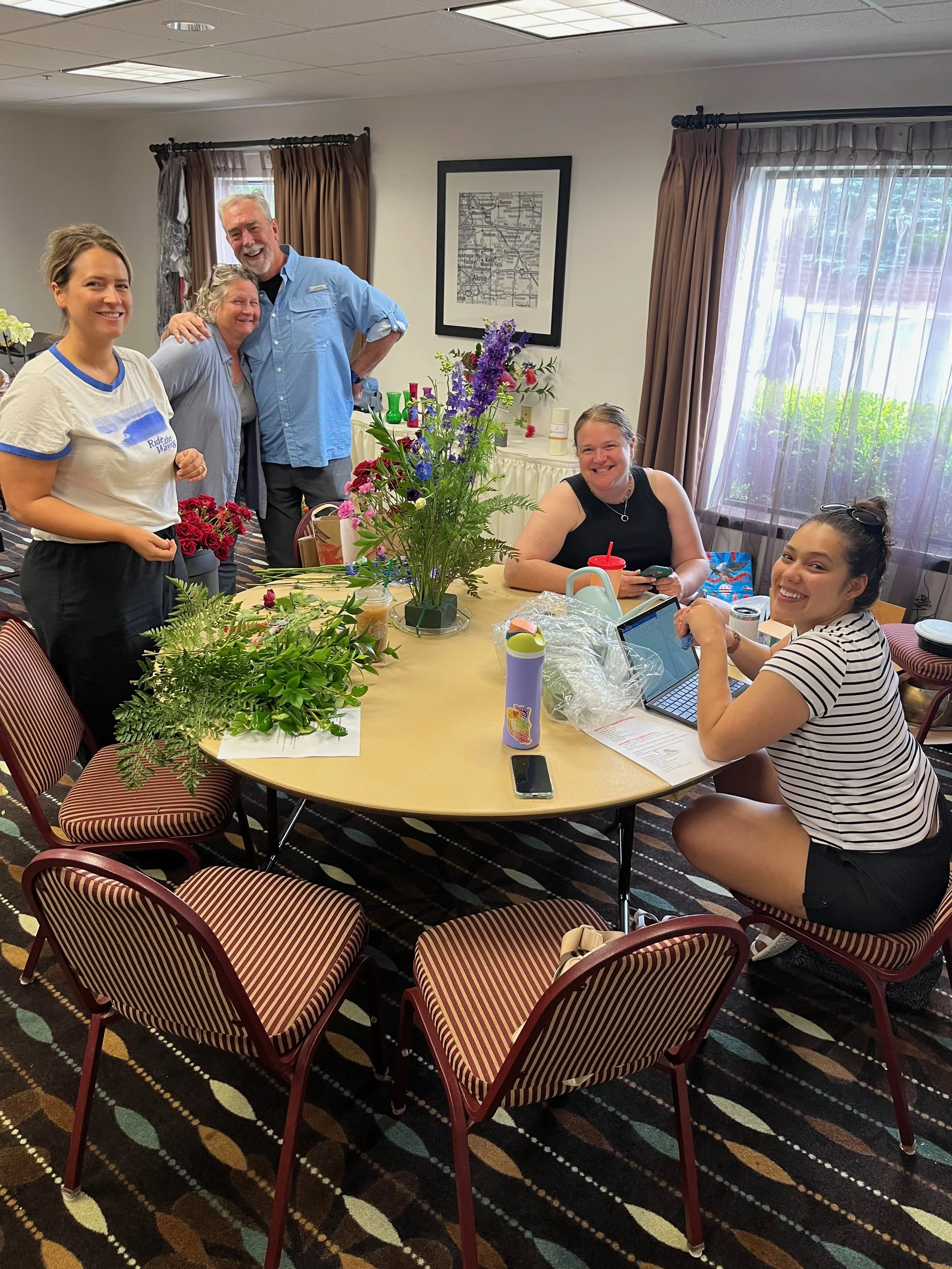 Five people in a room around a round table, some sitting and some standing, smiling, with flowers and beverages on the table, and a window with curtains in the background.