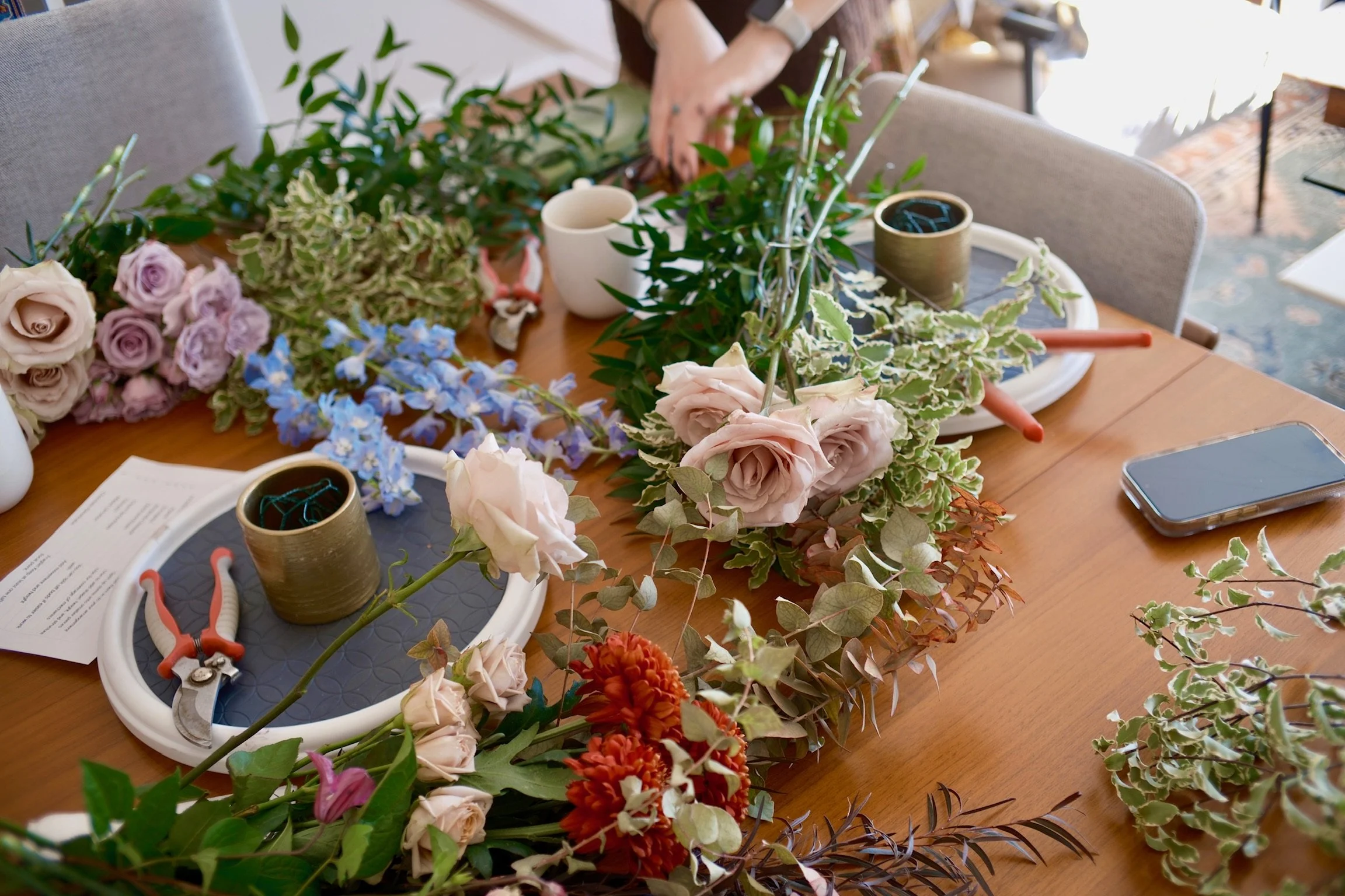 A table with various flowers and green foliage, some arranged and some loose, along with gardening tools, a smartphone, a paper, and cups.