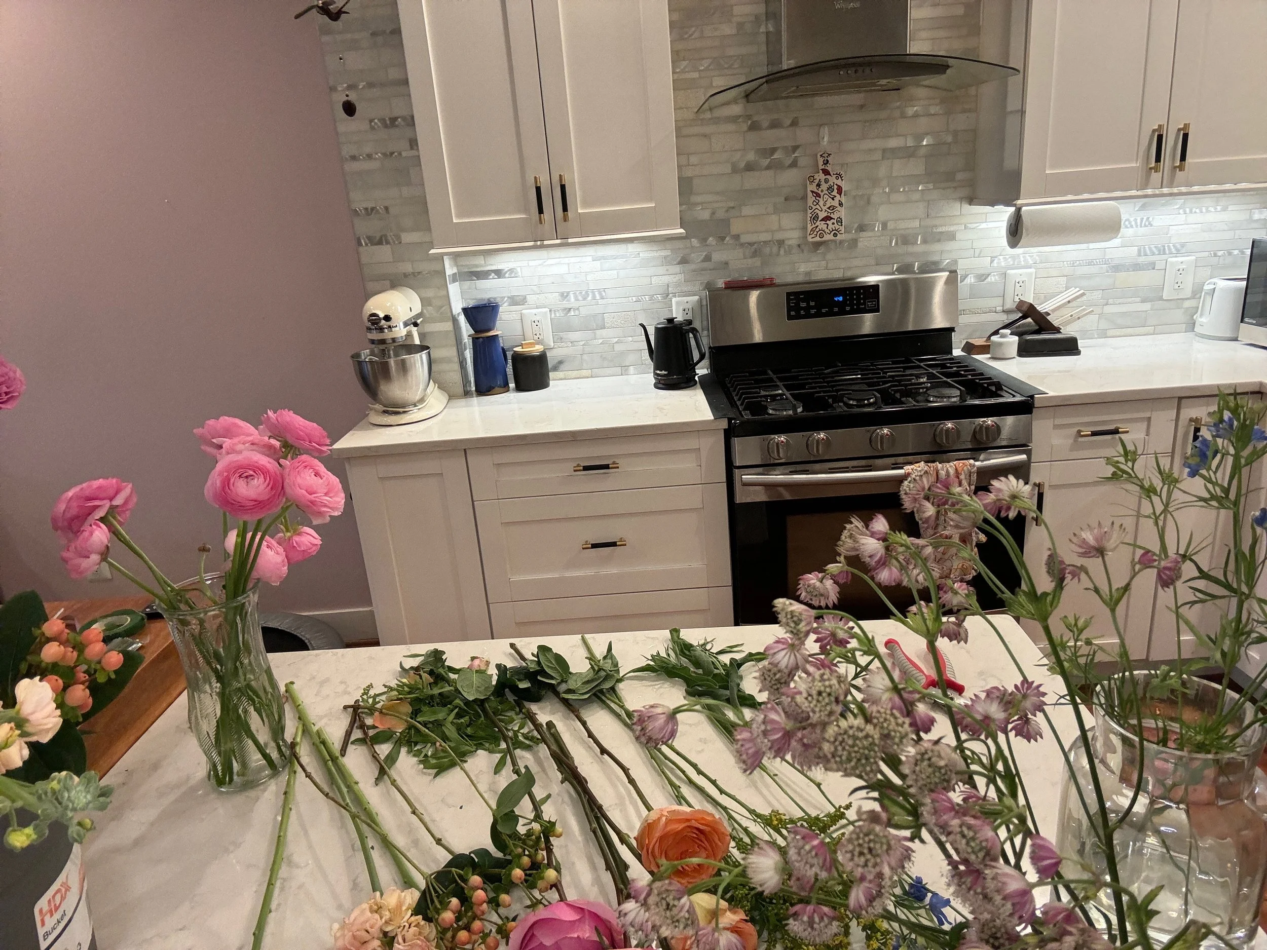 A modern kitchen with white cabinets, a grey tiled backsplash, and a black stove. The kitchen counter has various items including a kettle, a paper towel roll, and a small black container. There are colorful flowers in vases and flower arrangements o