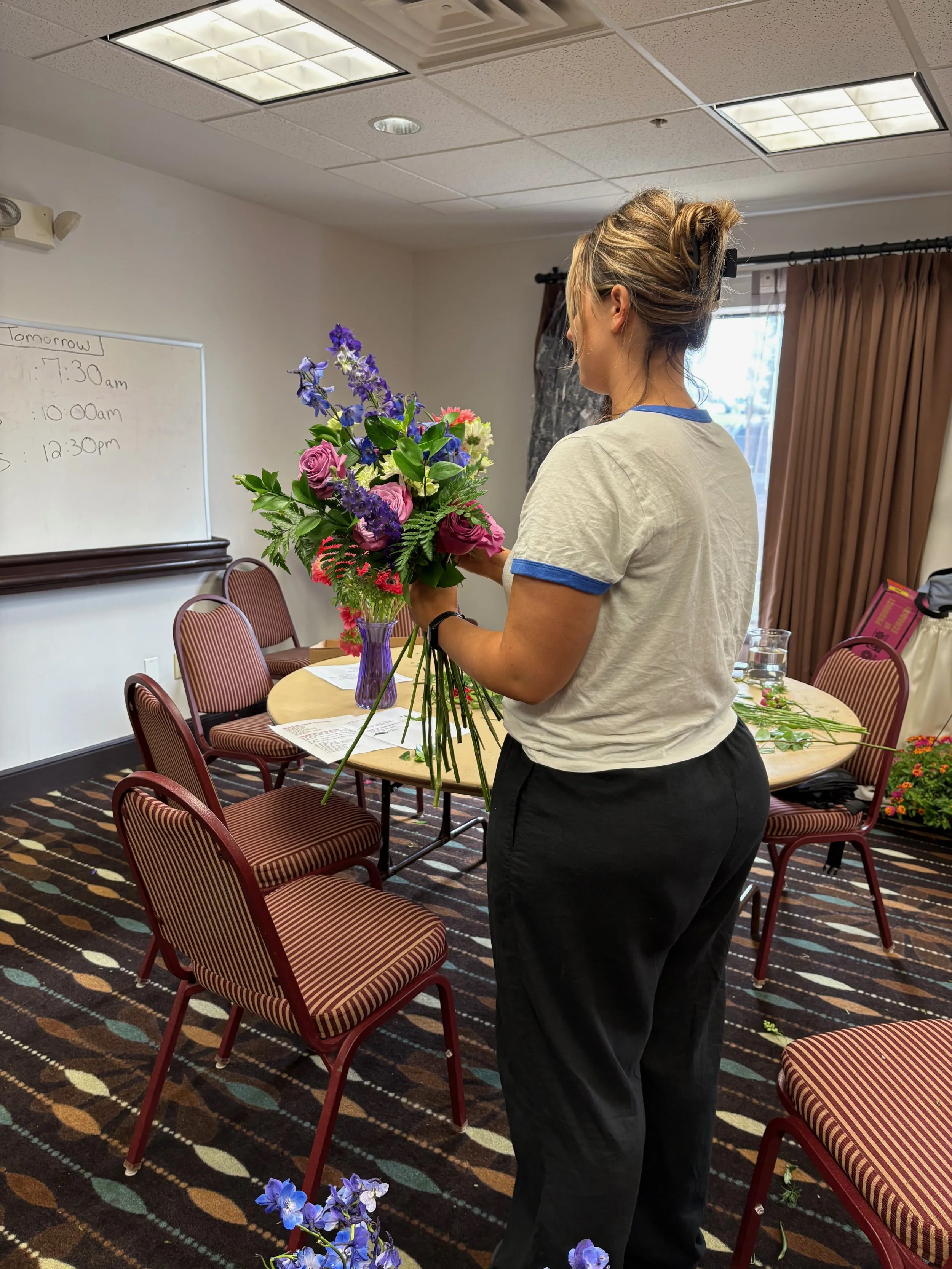 A woman arranging a bouquet of colorful flowers in a purple vase inside a conference room with red and beige chairs and a whiteboard.