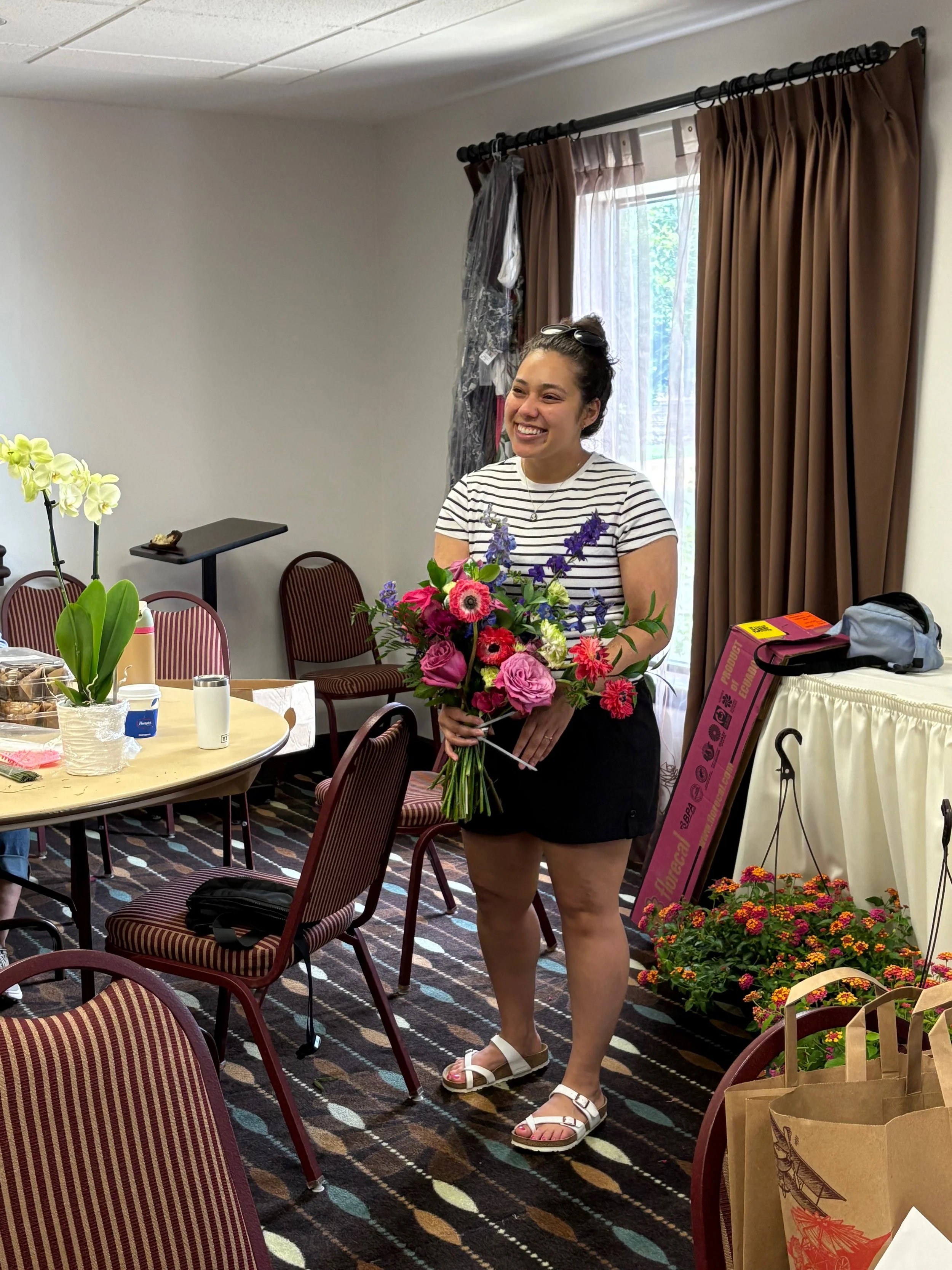 A woman holding a bouquet of flowers, smiling, wearing a striped t-shirt, black shorts, and sandals, standing in a room decorated with plants, chairs, and a window with brown curtains.