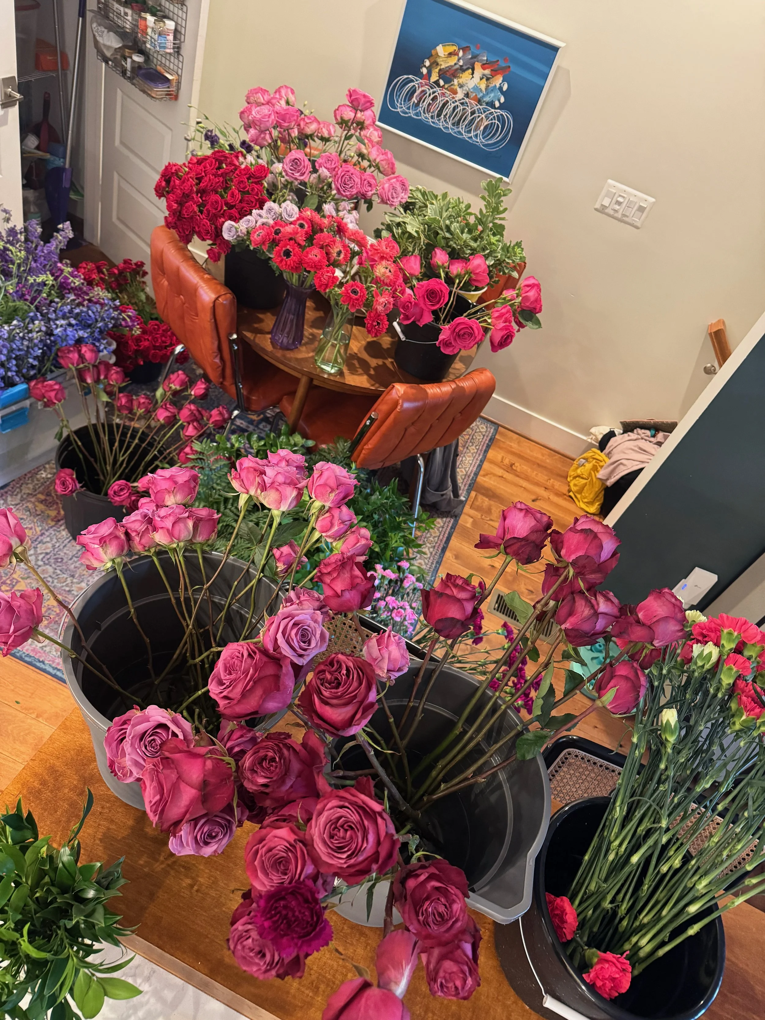 A room filled with various arrangements of pink and red roses and other flowers on a wooden table, with chairs and wall art in the background.