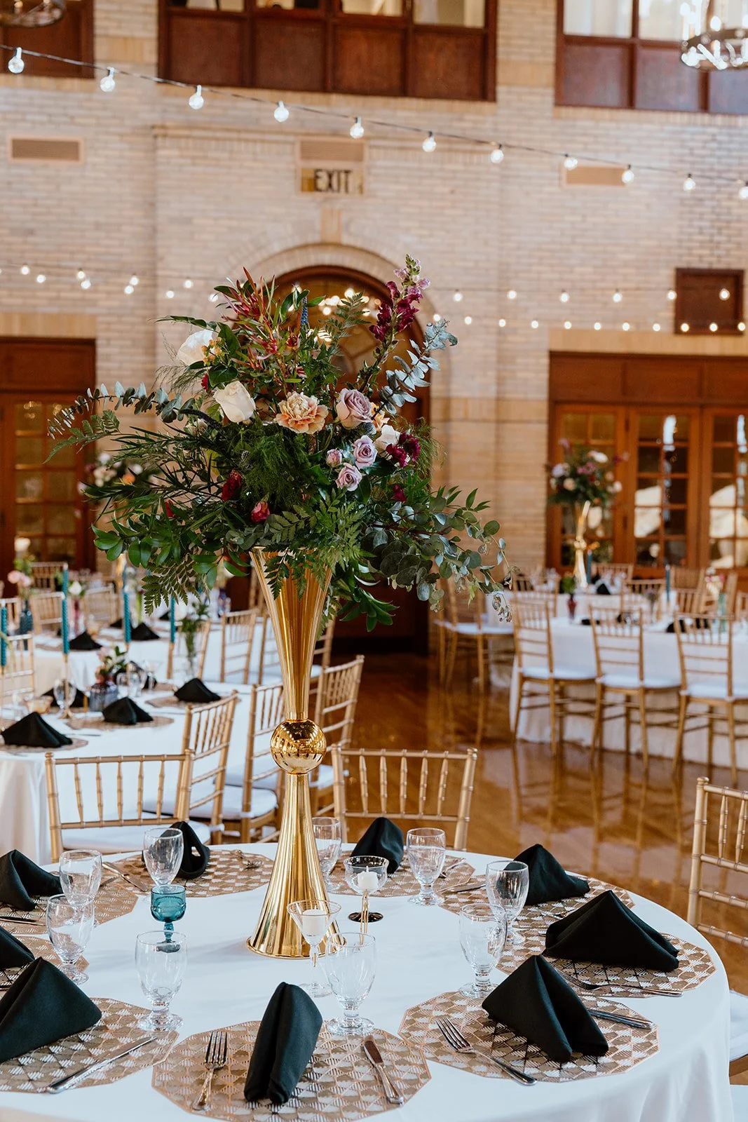 A table set for a formal event with a tall floral centerpiece in a gold vase, black napkins, wine glasses, and cutlery, in a setting with wood chairs and a brick wall with string lights.