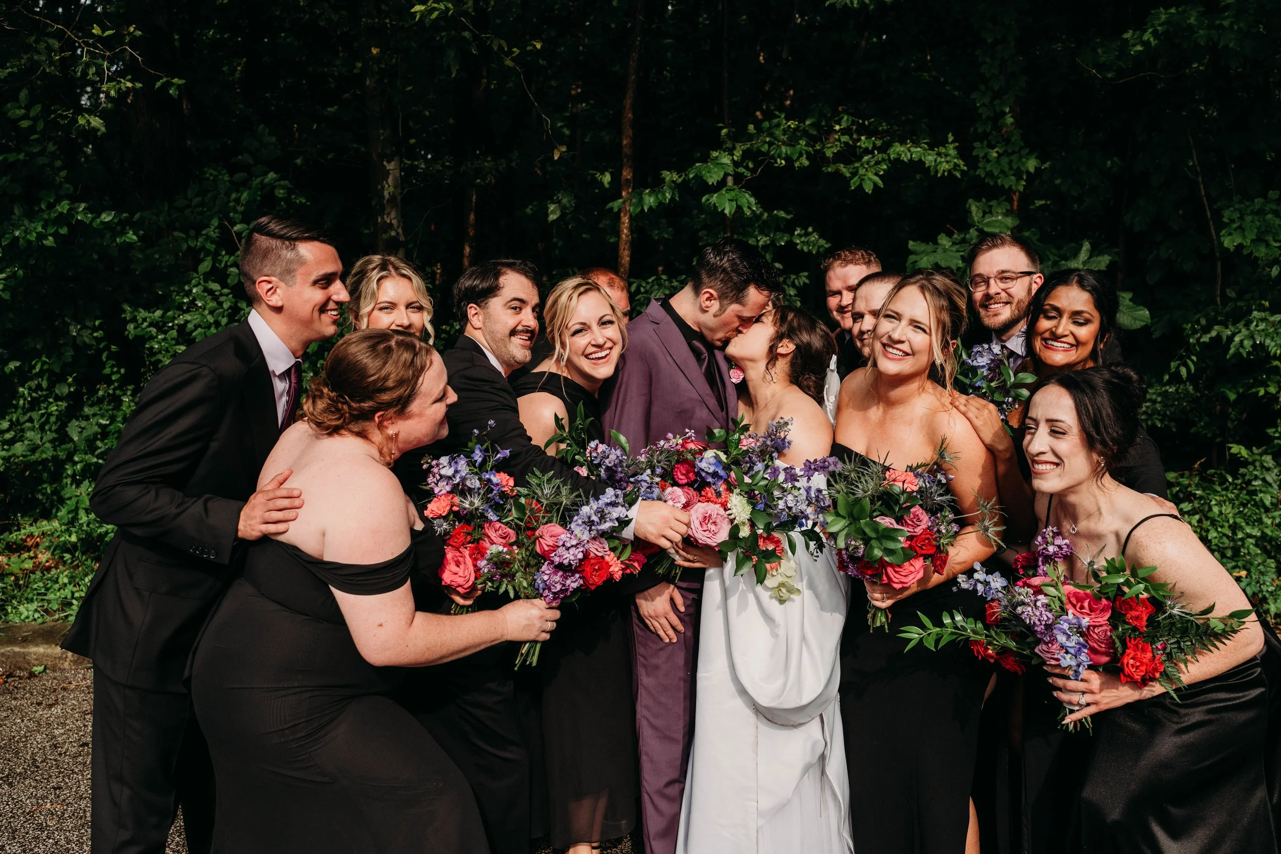 Bridesmaids and groomsmen holding bouquets of red, purple, blue, pink, and green flowers huddle around a bride and groom kissing. 