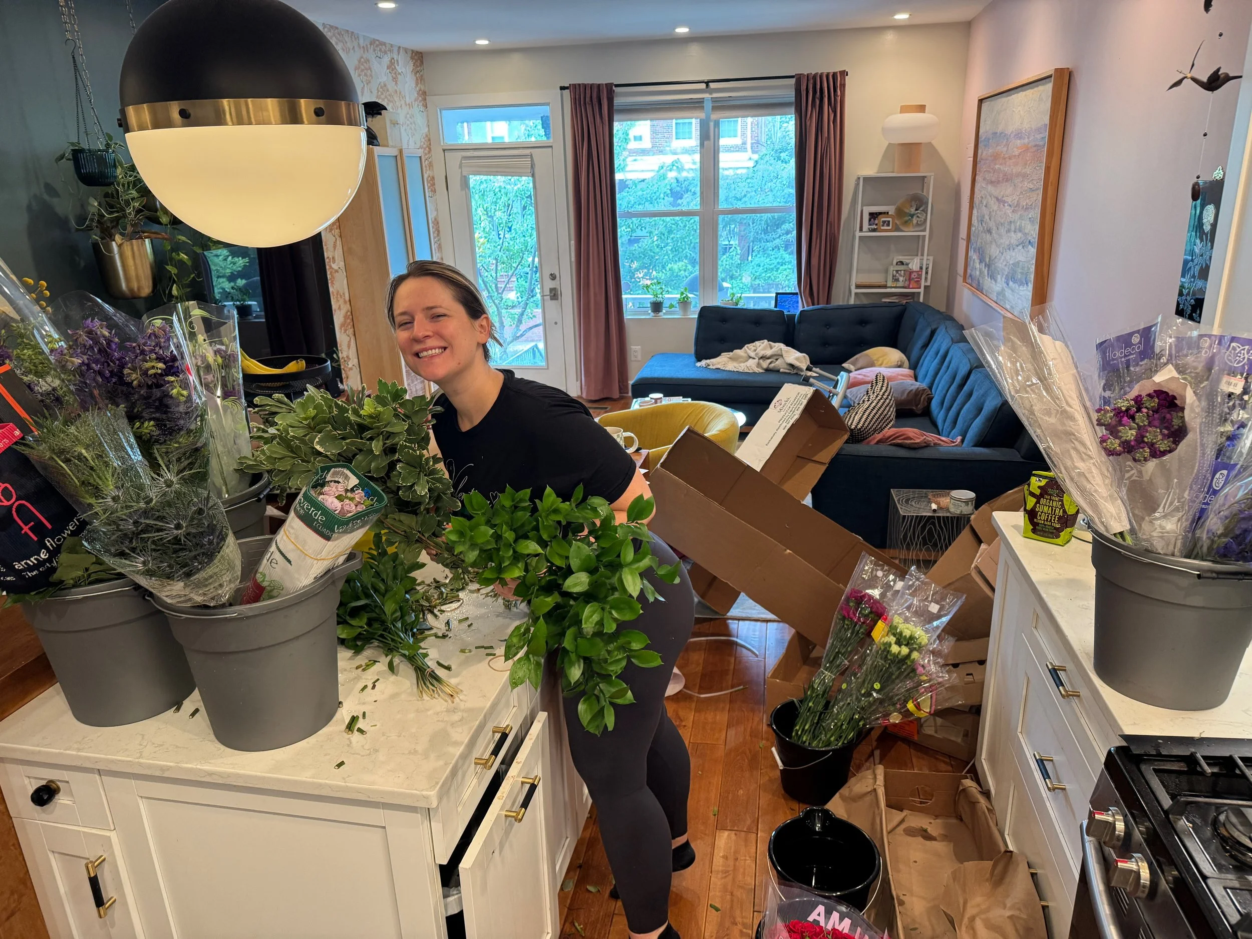A woman smiling in a kitchen surrounded by flower arrangements, boxes, and packing materials, with a living room in the background.