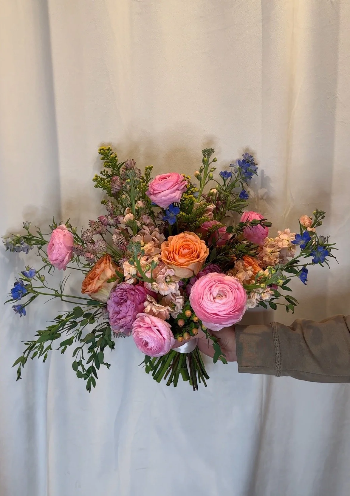 A hand holding a colorful bouquet of pink, orange, purple, and blue flowers against a plain background.