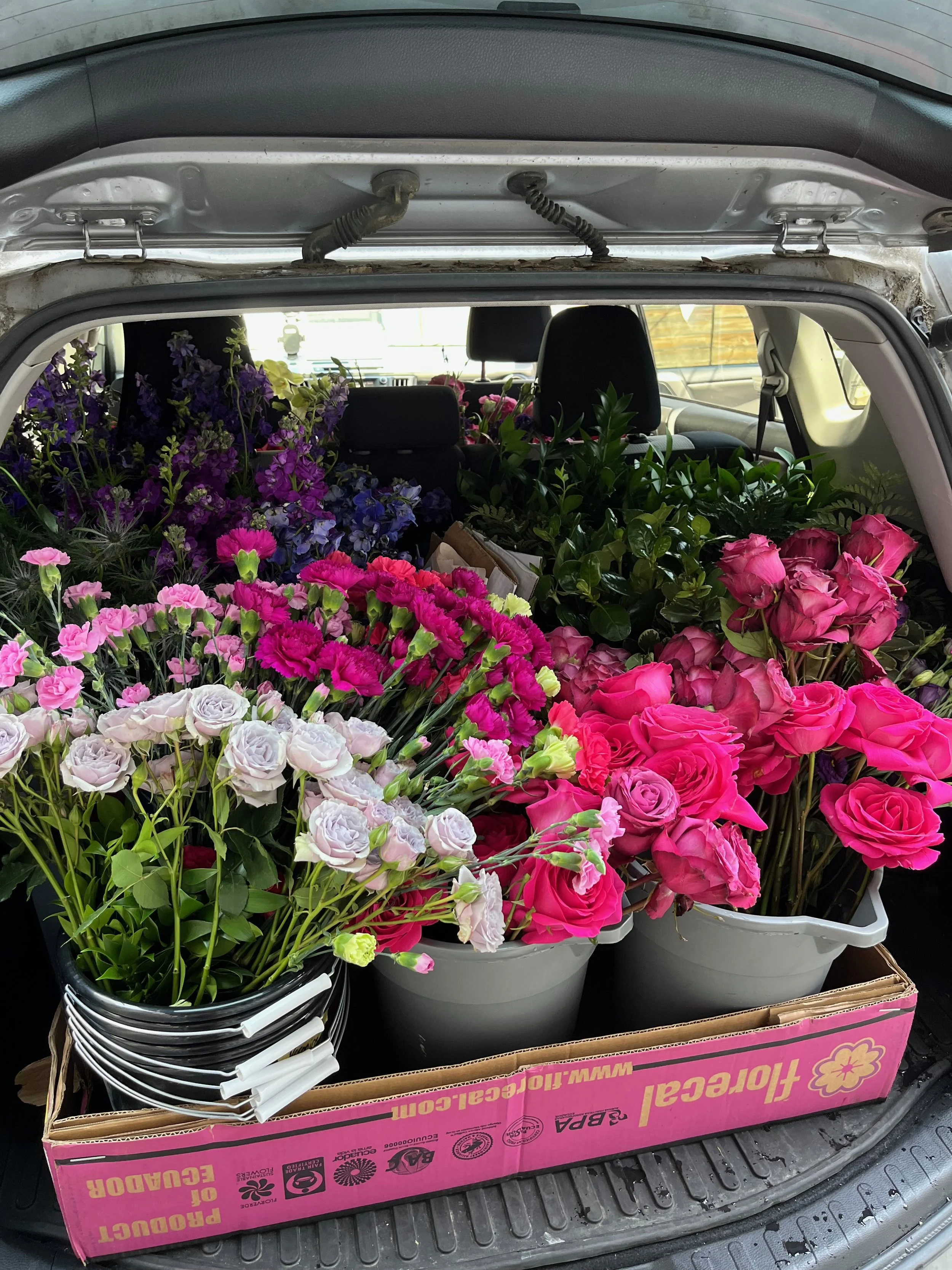 The back of a car filled with various colorful flowers, including pink roses, purple and blue flowers, in a pink and white cardboard box.