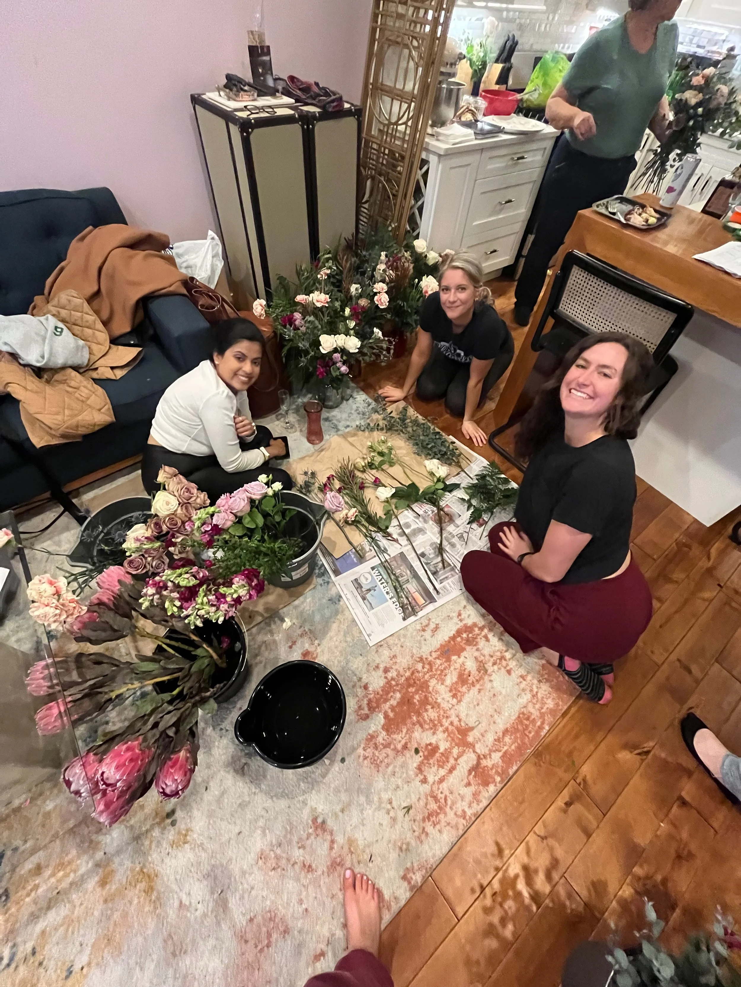 Four women arranging flowers on a table in a living room, surrounded by various gardening supplies, with a person in the background holding a bouquet of flowers.