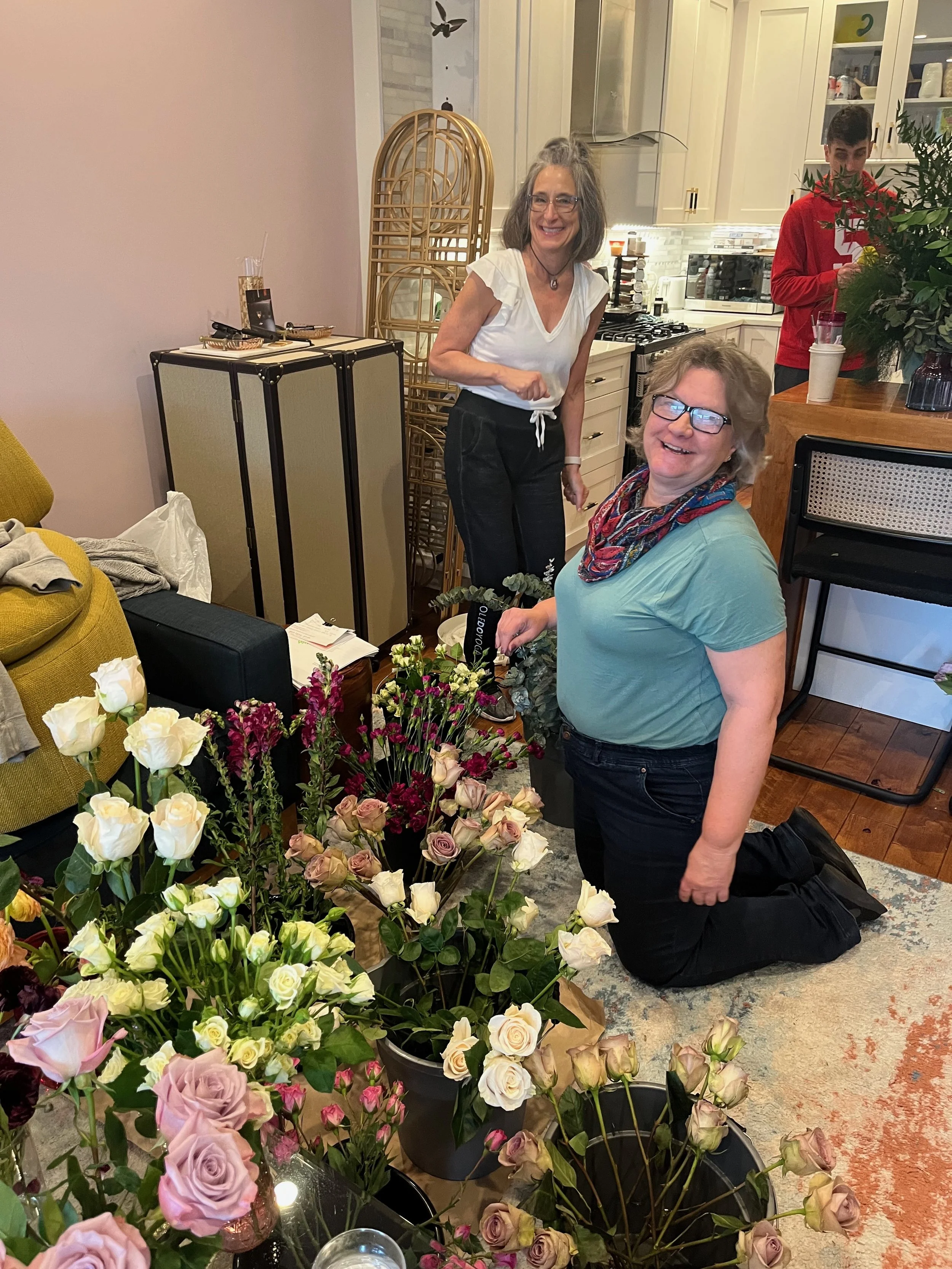 Two women are surrounded by various flower arrangements in a kitchen. One is kneeling, smiling, wearing glasses, a teal shirt, and black pants with a colorful scarf. The other is standing, smiling, wearing a white top and dark pants. There is a man i