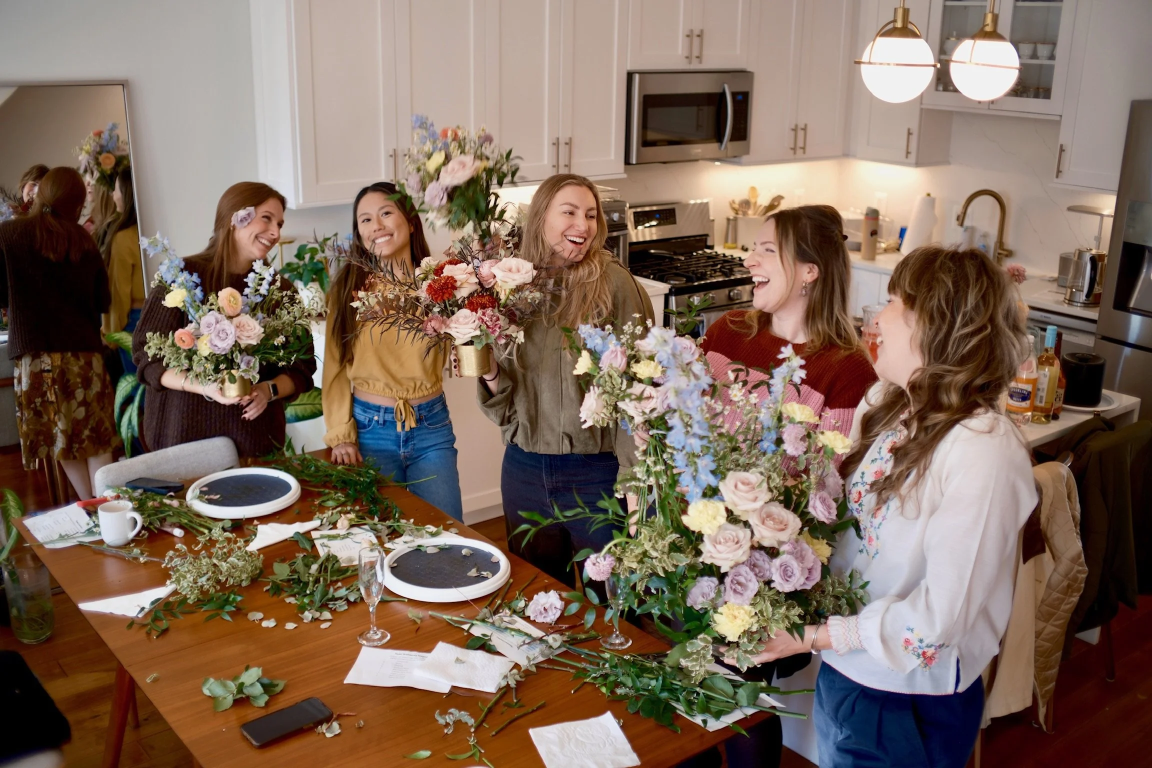 A group of women gathered in a kitchen, joyfully exchanging flowers and smiling, with floral arrangements and flower arranging supplies on a table.