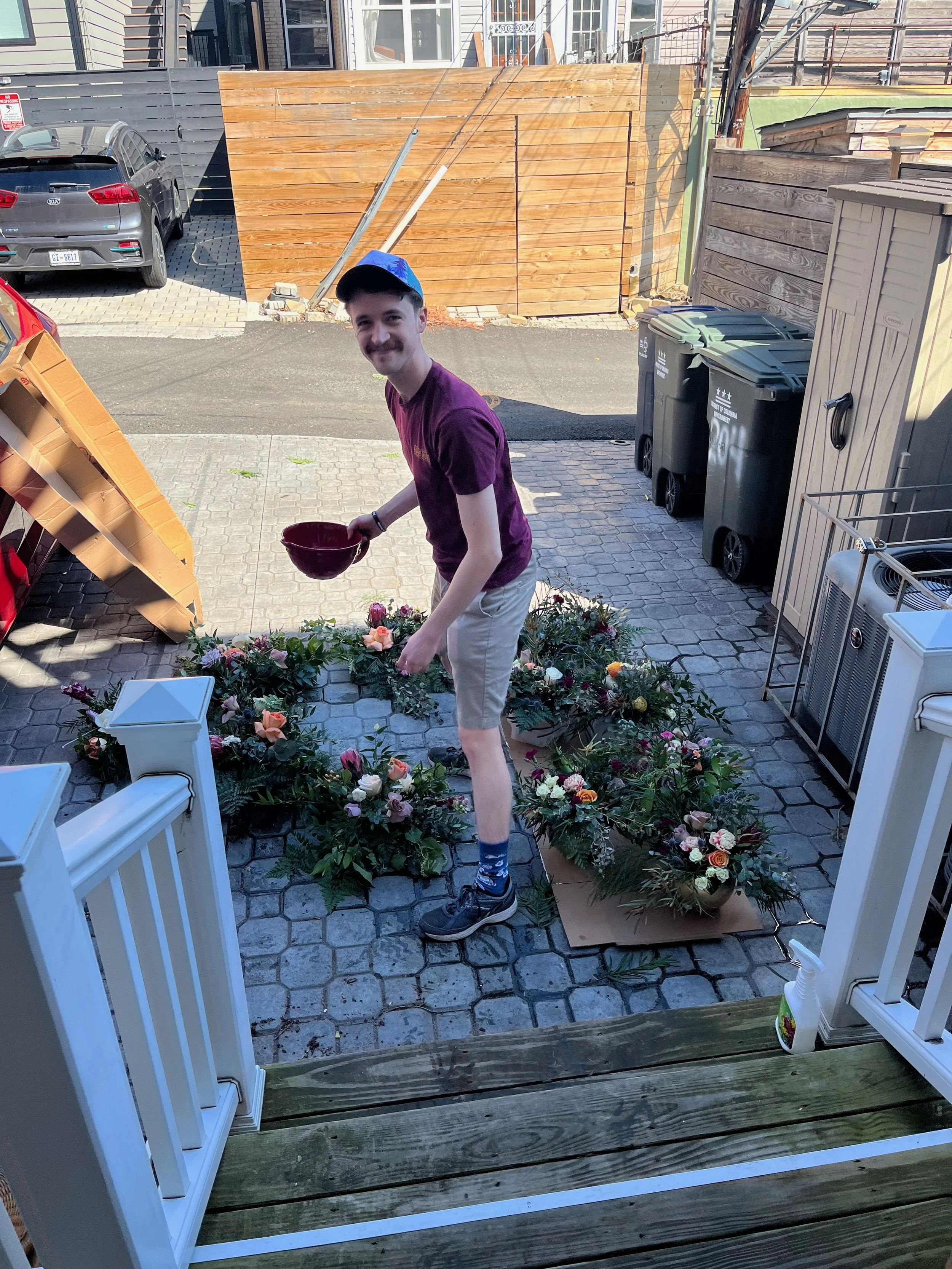 A young man arranging flowers on the porch in a circle, holding a red bowl, smiling, wearing a purple T-shirt, beige shorts, black sneakers, and a blue cap, surrounded by flower arrangements and greenery.