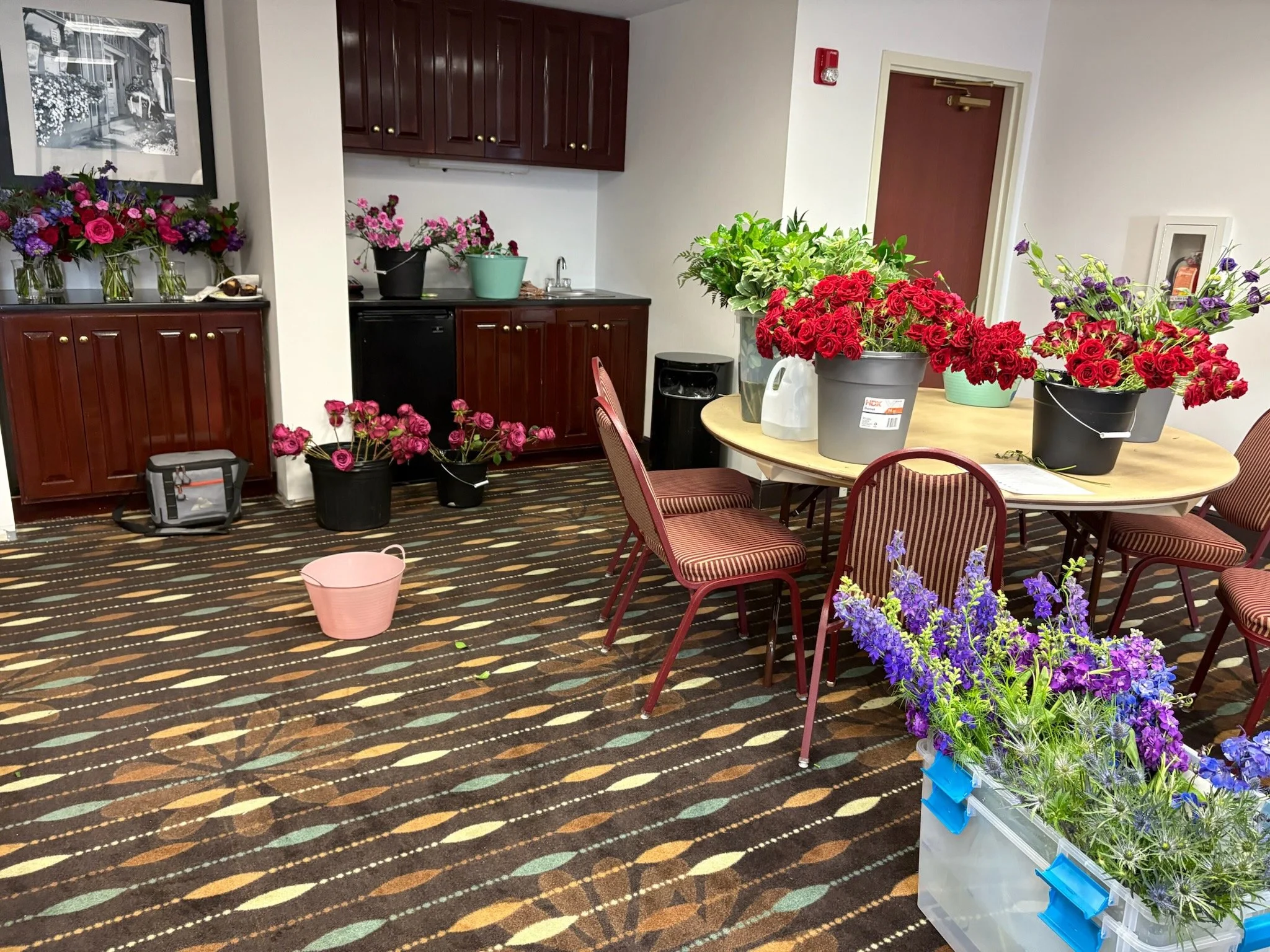 A room filled with colorful flowers on a round table, on the floor, and on shelves; chairs around the table; cabinets and framed pictures on the wall.
