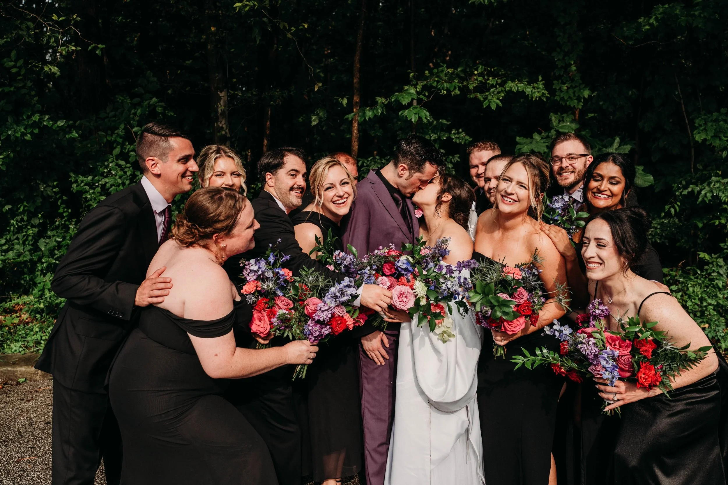 A wedding celebration with the bride and groom kissing surrounded by their wedding party holding bouquets, outdoors with greenery in the background.
