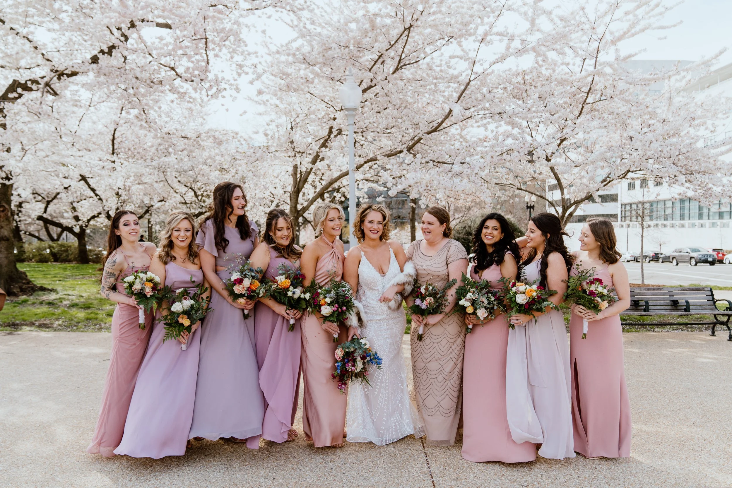 Bridesmaids wearing shades of pink and purple holding bouquets with lush greenery and many colored flowers surround a smiling bride in front of a blossoming cherry blossom tree.