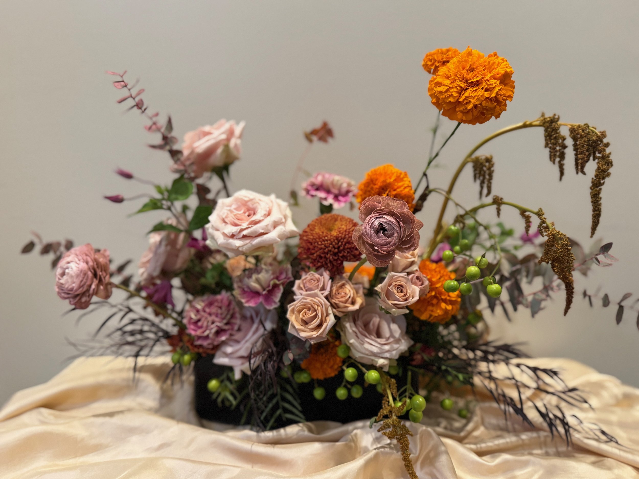 A floral arrangement in a black vase with pink roses, orange marigolds, and purple ranunculus, along with various greenery, placed on a cream-colored satin cloth against a plain white background.