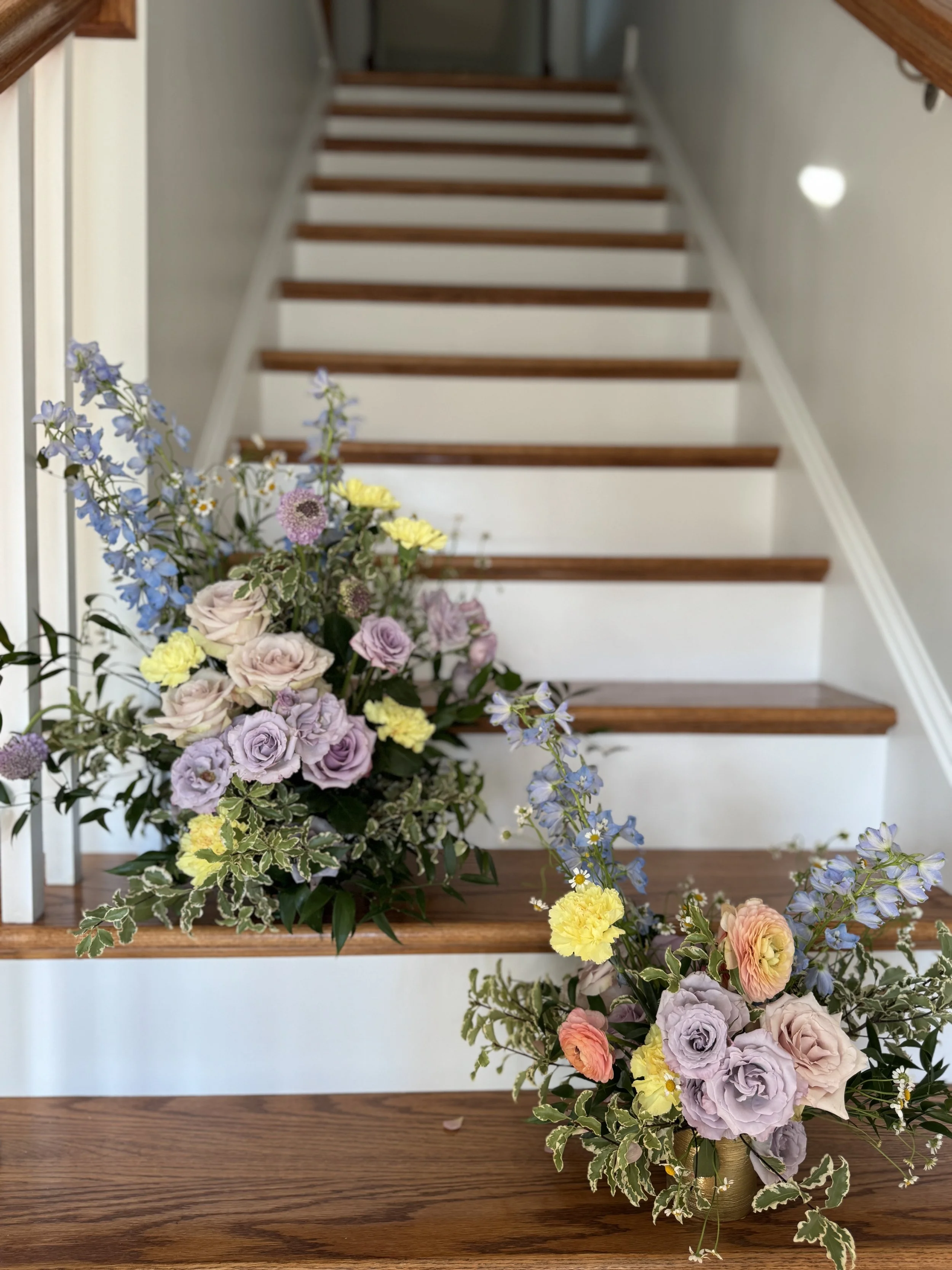A floral arrangement with purple, yellow, peach, and pink flowers on a wooden table in front of a staircase with white risers and wooden treads.