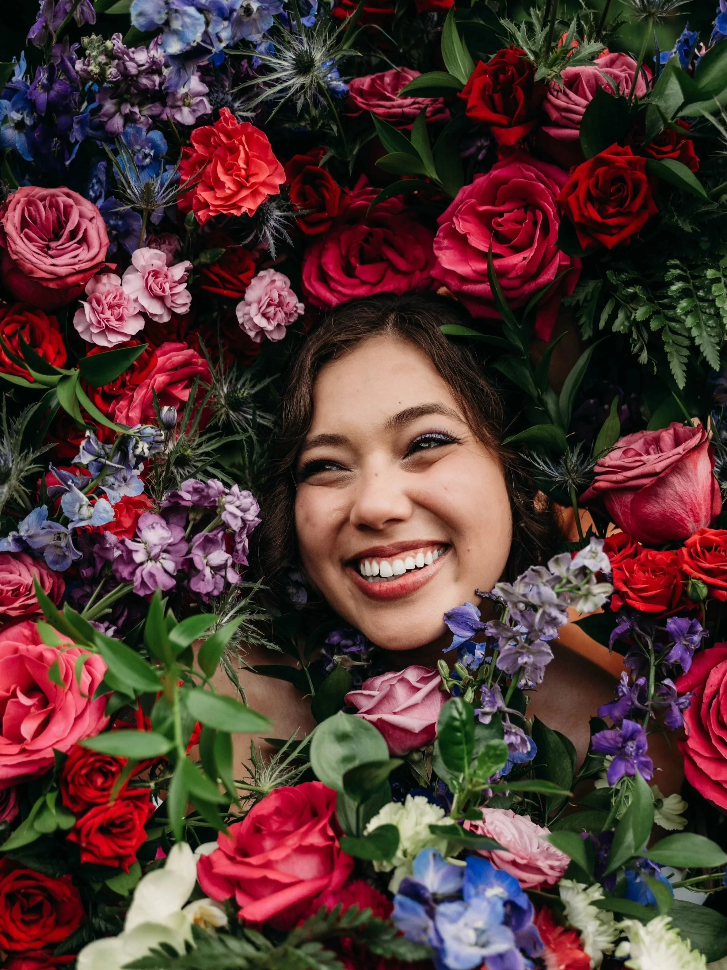 A woman smiling and lying amidst colorful flowers including roses and other blooms.