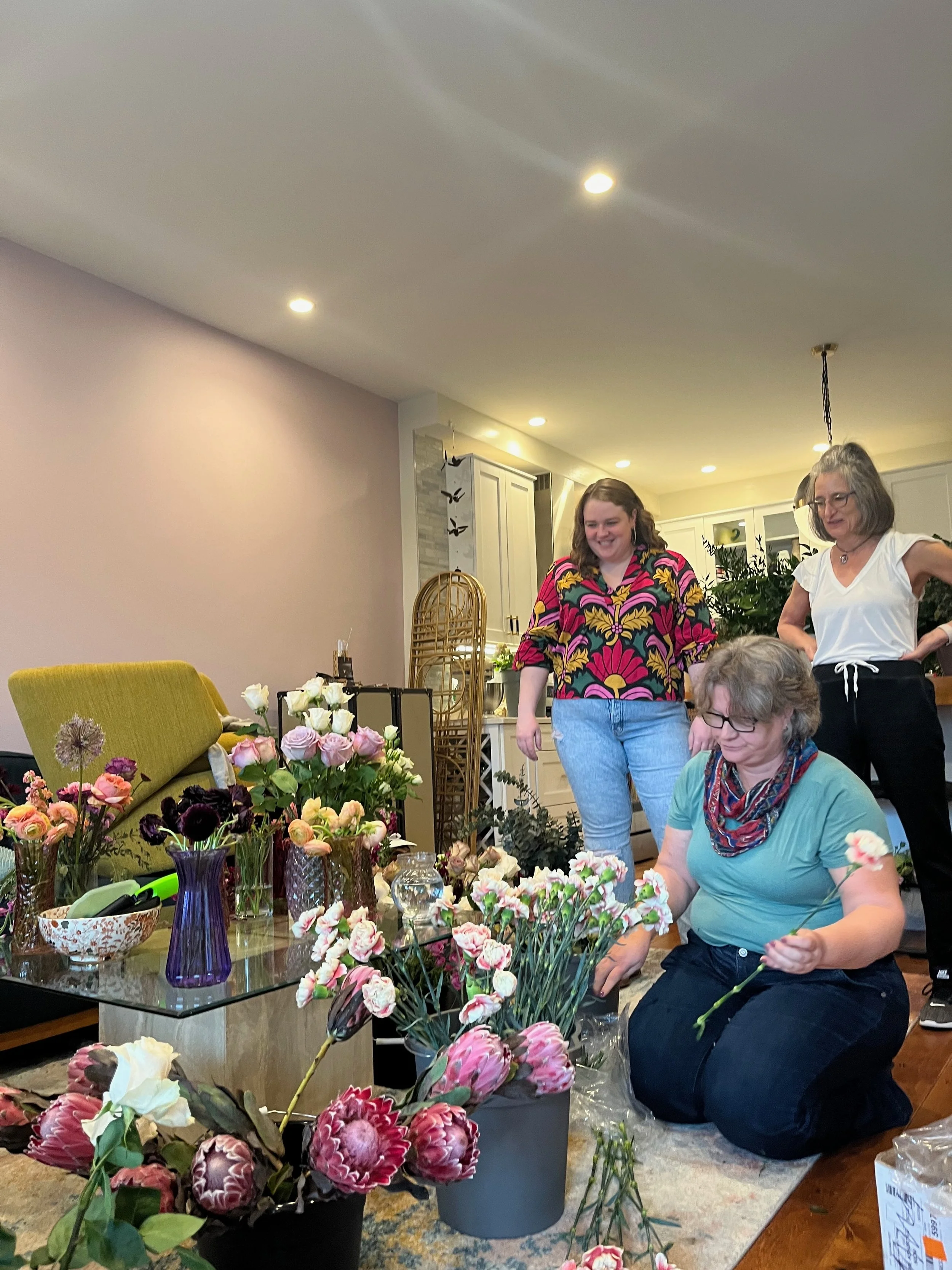 Three women arranging flowers in a living room with a coffee table of floral arrangements and vases.