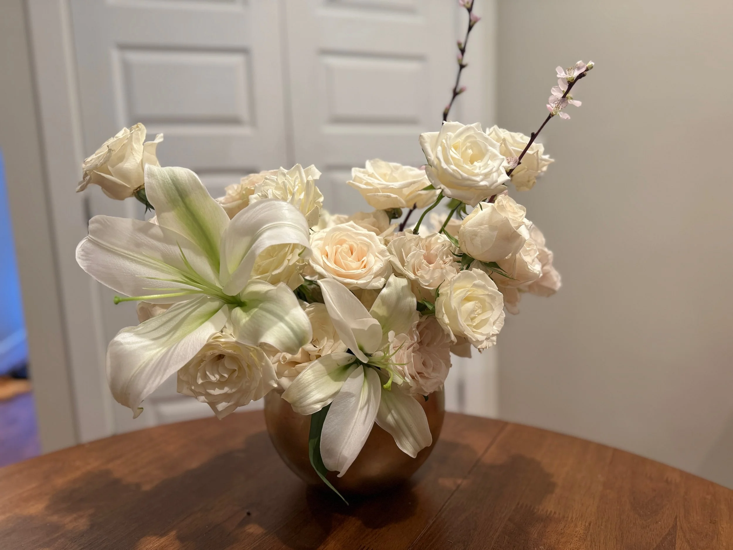 A floral arrangement in a gold vase on a wooden table, featuring white lilies, white roses, and pink cherry blossoms.