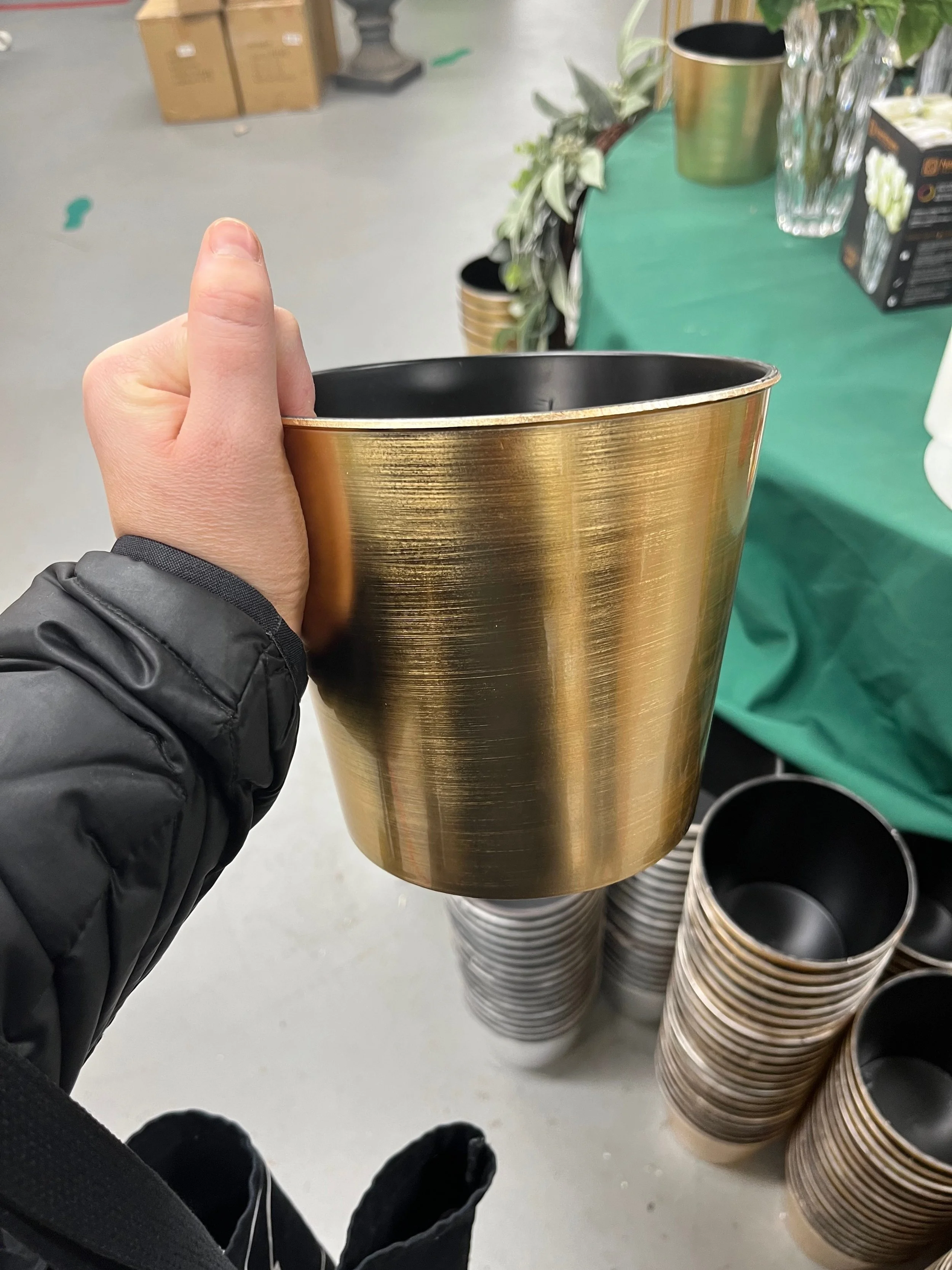 A person holding a gold-colored metallic cup, with several similar cups stacked on a display in the background at a store or market.