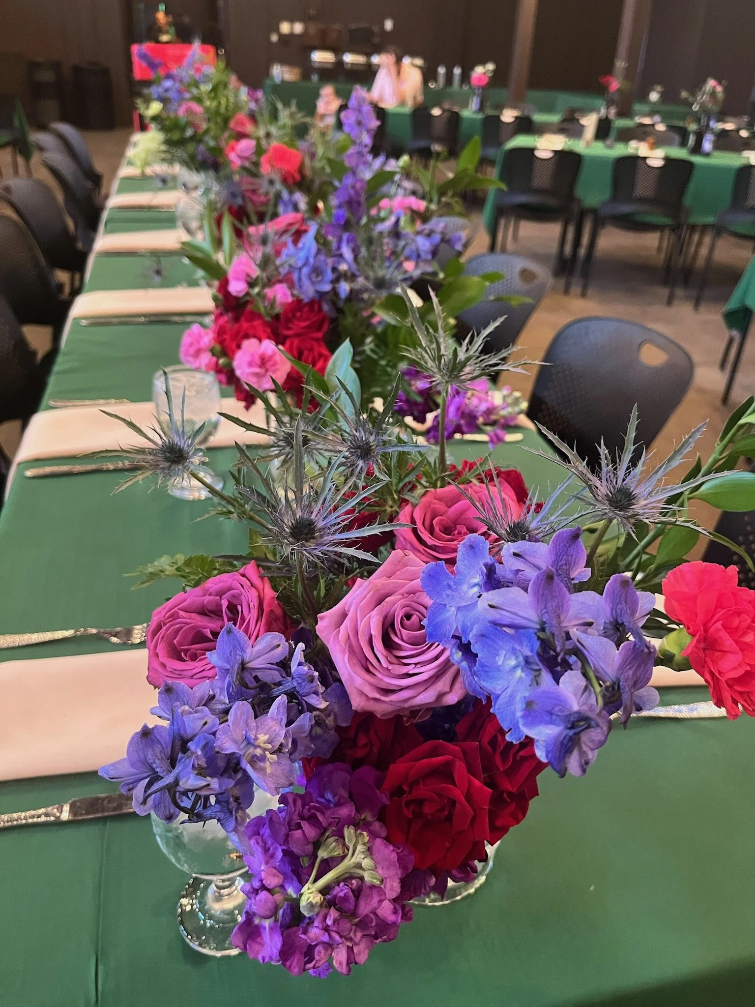 Colorful floral centerpiece with pink roses, purple delphiniums, red carnations, and thistle, placed on a green tablecloth in a banquet hall.